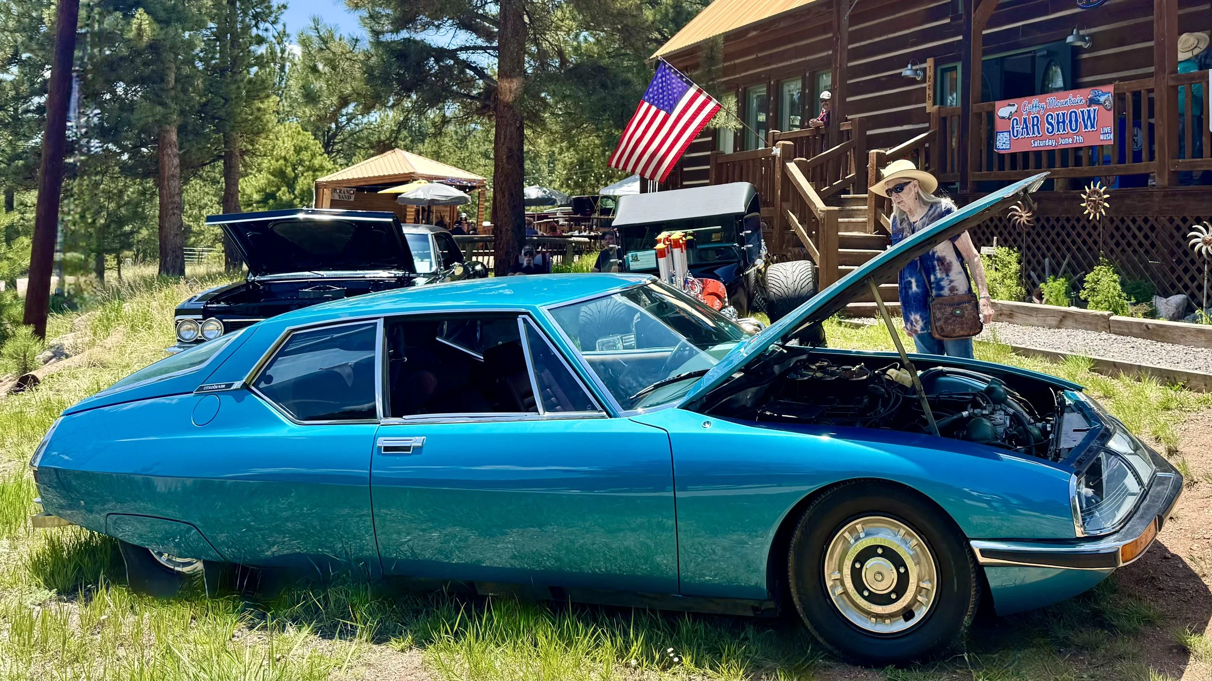 Blue vintage sports car with open hood on grassy field at car show, woman in sun hat and sunglasses examining it, other classic cars and trees in background, American flag and signs indicating car show event.