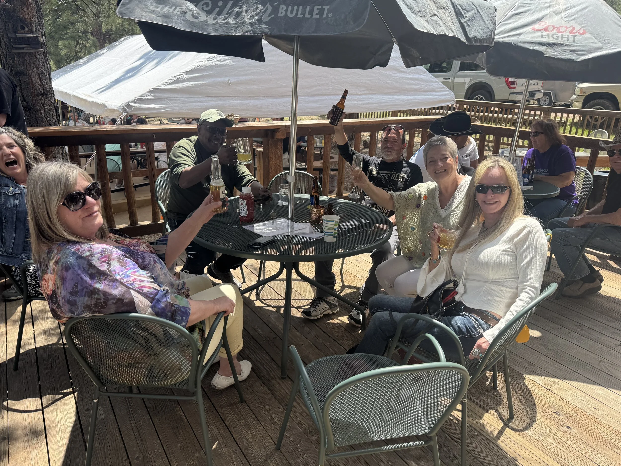 Group of people sitting at a round outdoor table on a wooden deck, enjoying drinks and socializing in sunlight, with umbrellas providing shade.