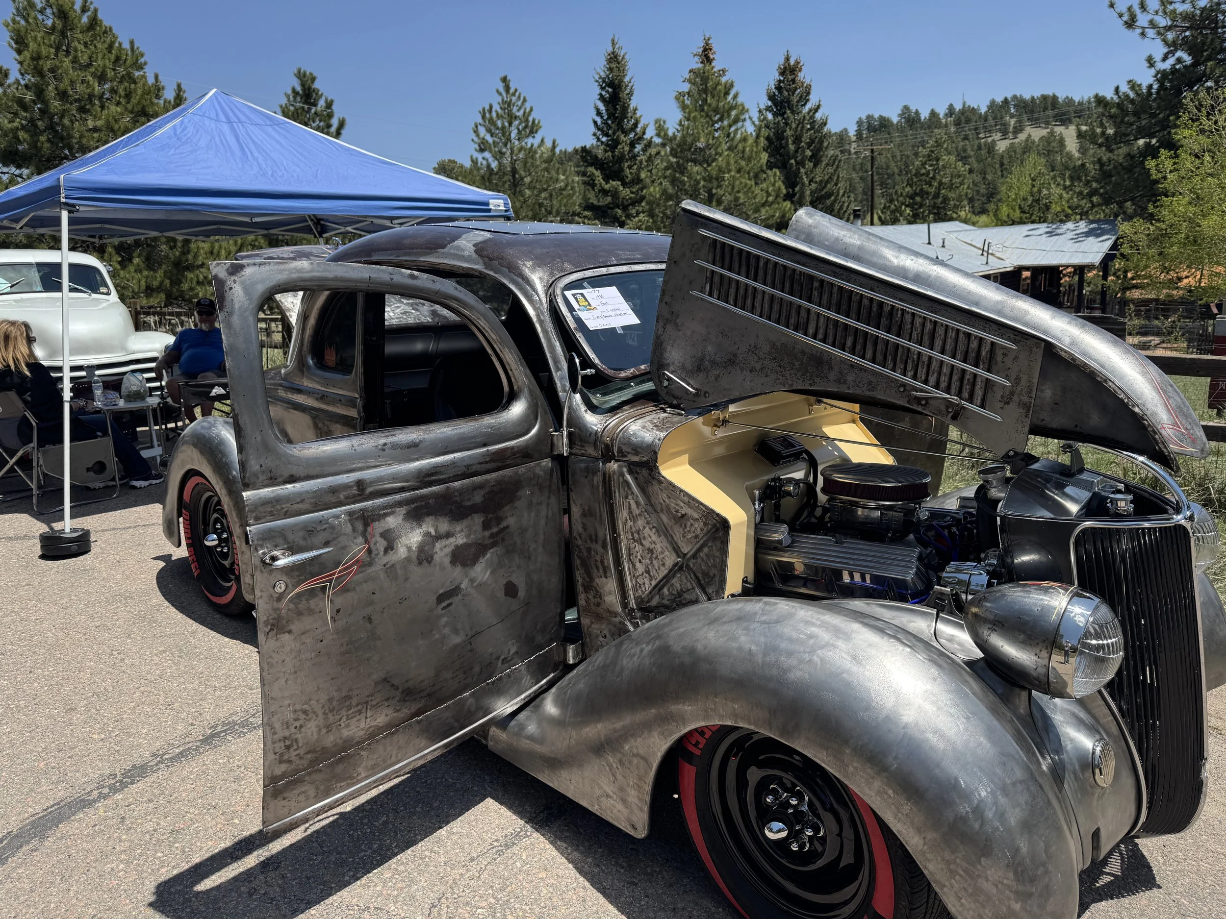 Vintage hot rod car with an exposed engine, parked outdoors at a car show with a blue canopy and several people sitting nearby under the canopy.