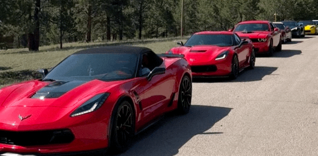 A line of five red sports cars parked on the side of a road with trees in the background.