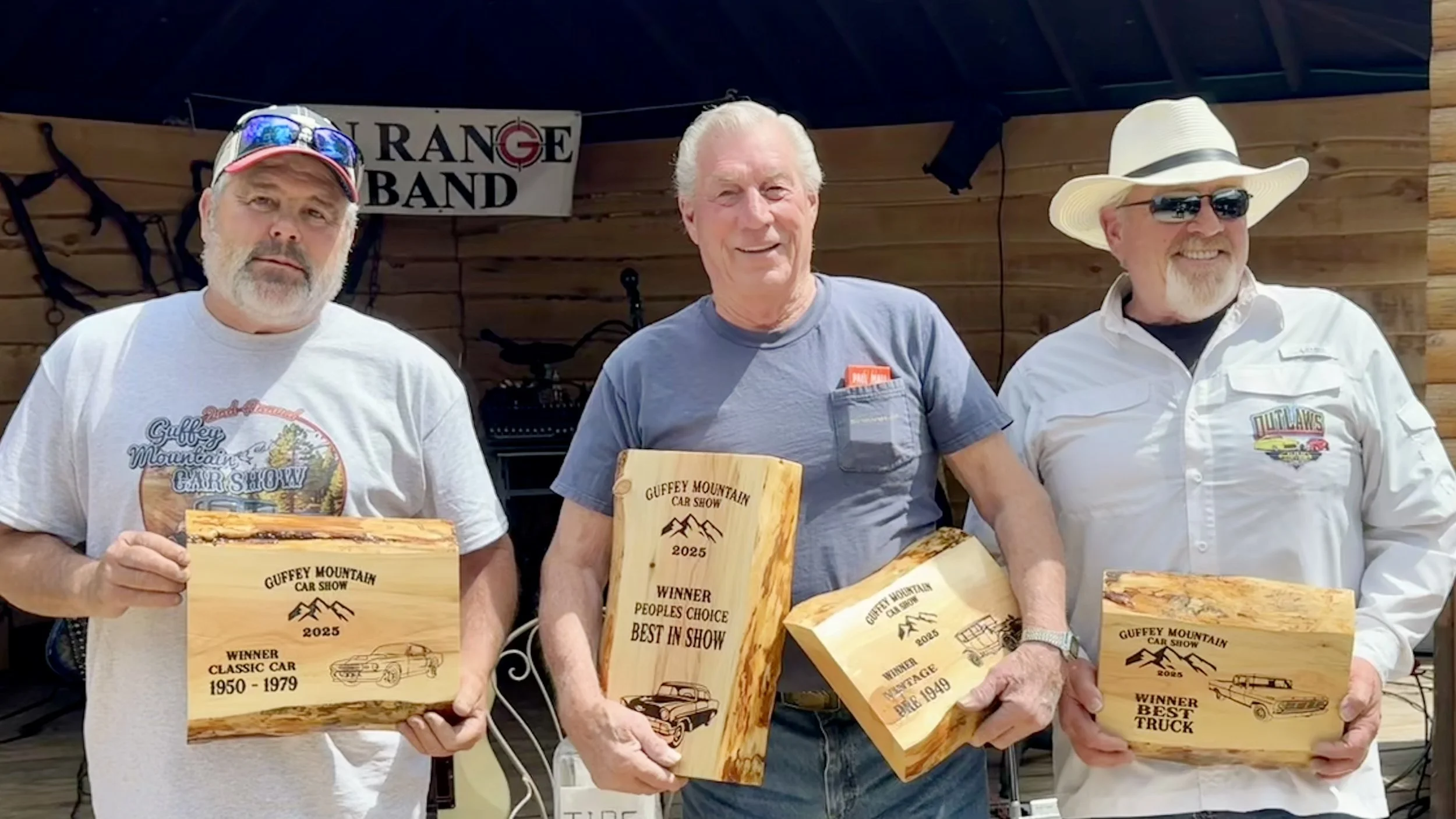 Three men are standing outdoors, each holding wooden awards from the Guffey Mountain Car Show, with a banner behind them reading 'Guffey Band'. The awards are engraved with car show titles and years.