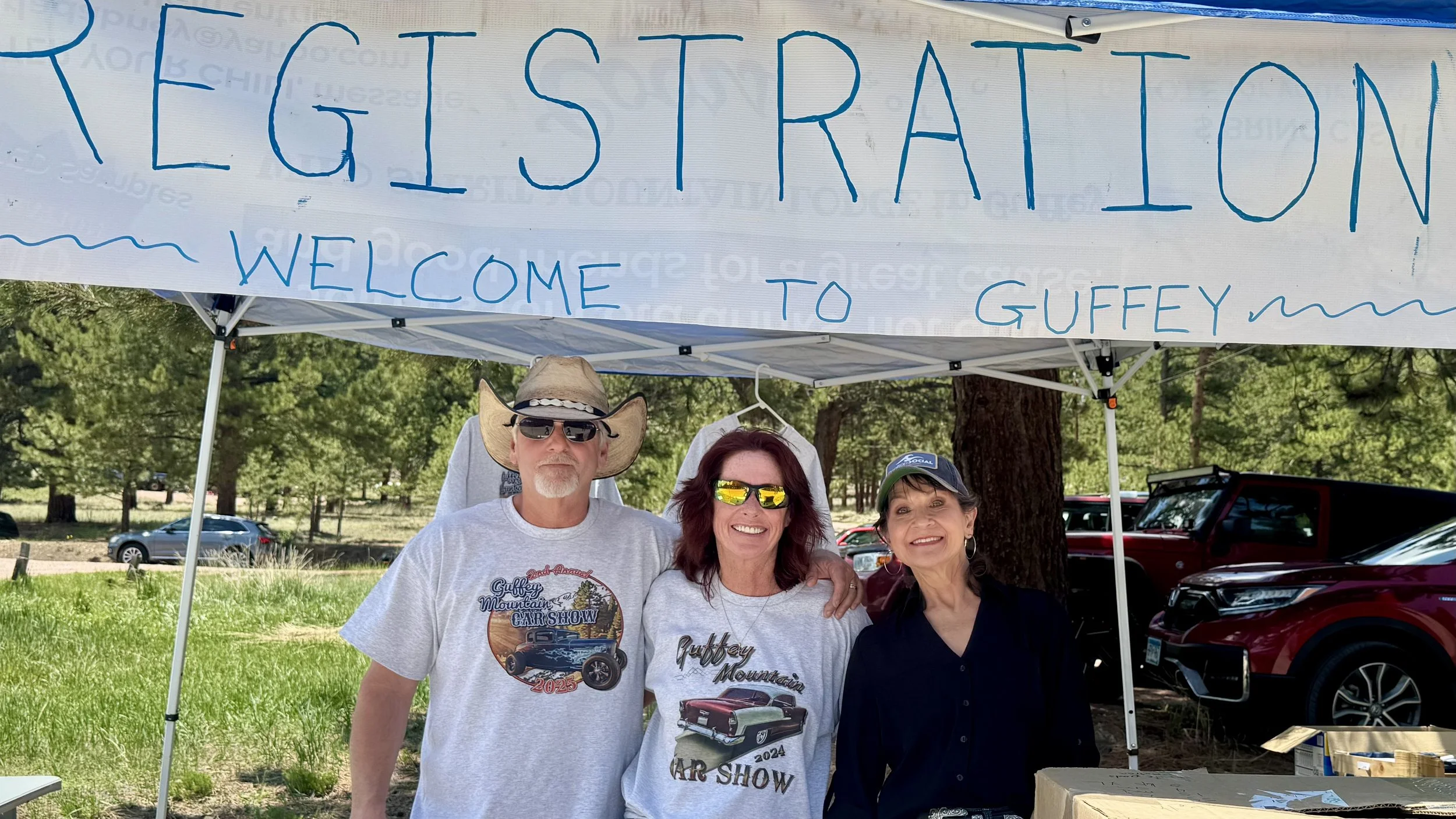 Three people standing under a tent with a sign that reads 'REGISTRATION WELCOME TO GUFFEY' at a car show event, outdoors on a sunny day with trees and parked cars in the background.