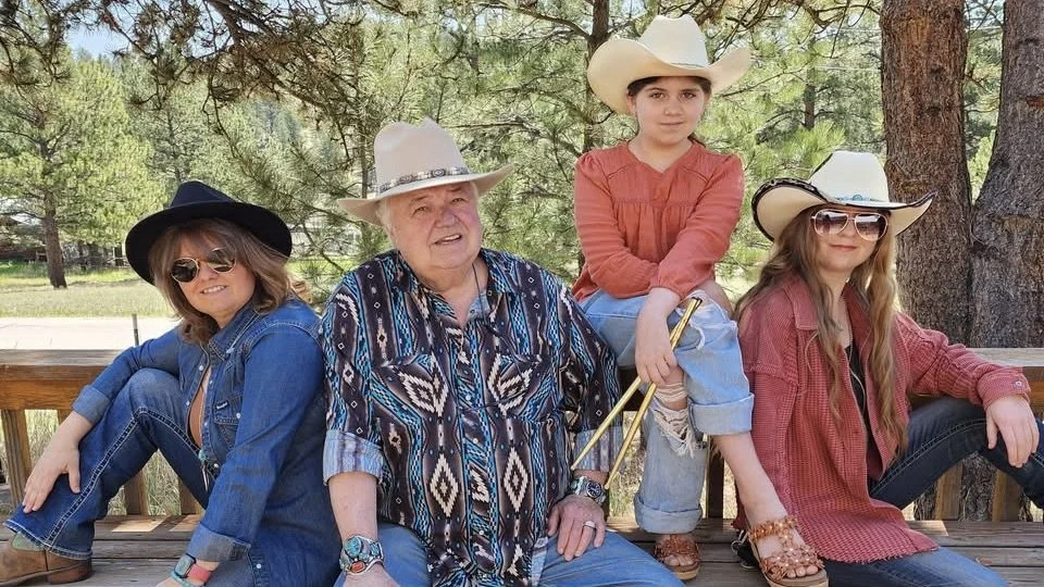 Four people, three women, and one man sitting on a wooden bench outdoors. They are wearing cowboy hats and casual clothing, with a background of trees and greenery.
