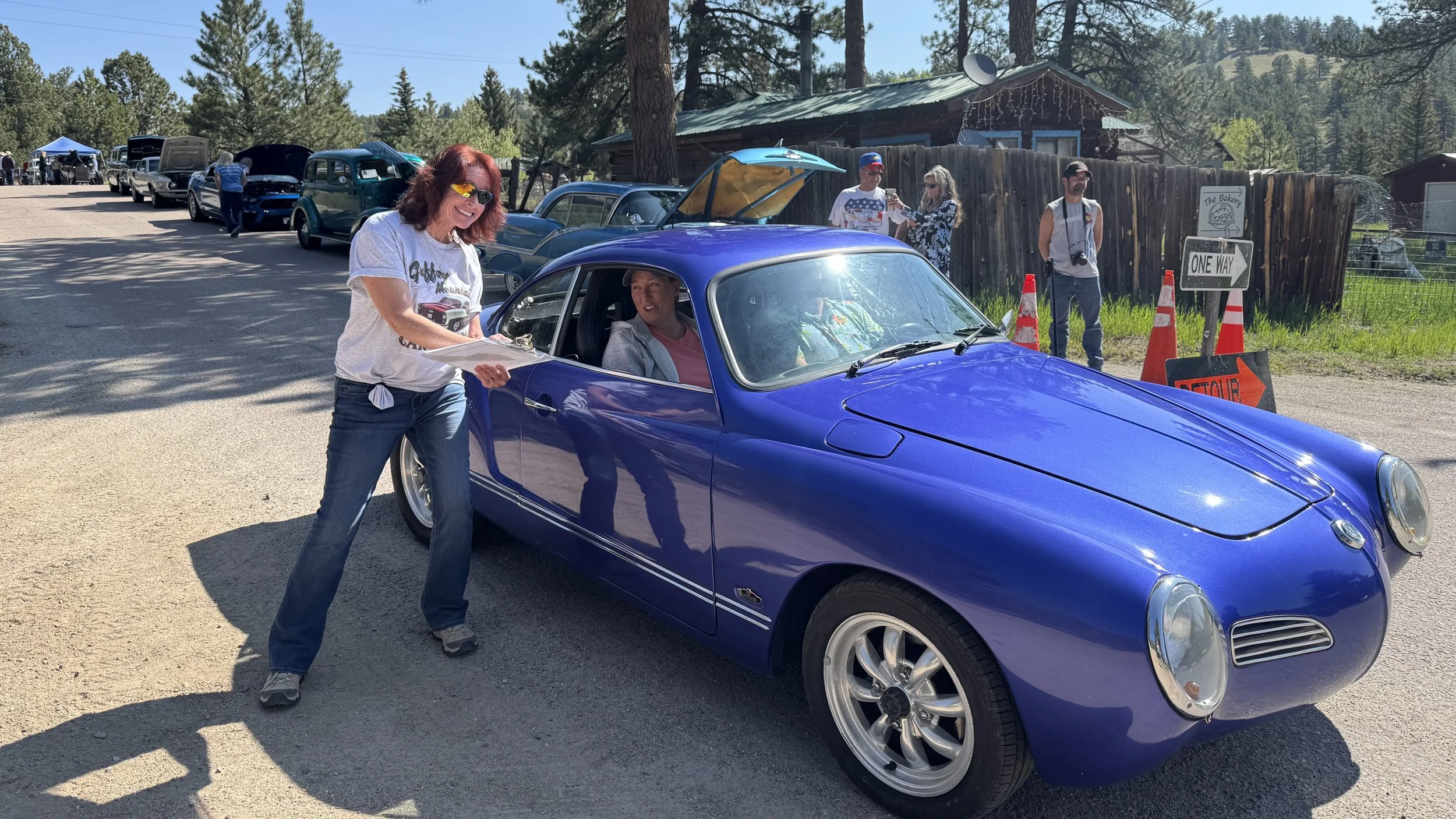 A woman with red hair and sunglasses is smiling and handing a clipboard to a man sitting inside a bright blue vintage car at a classic car show. Several other classic cars and people are visible in the background.