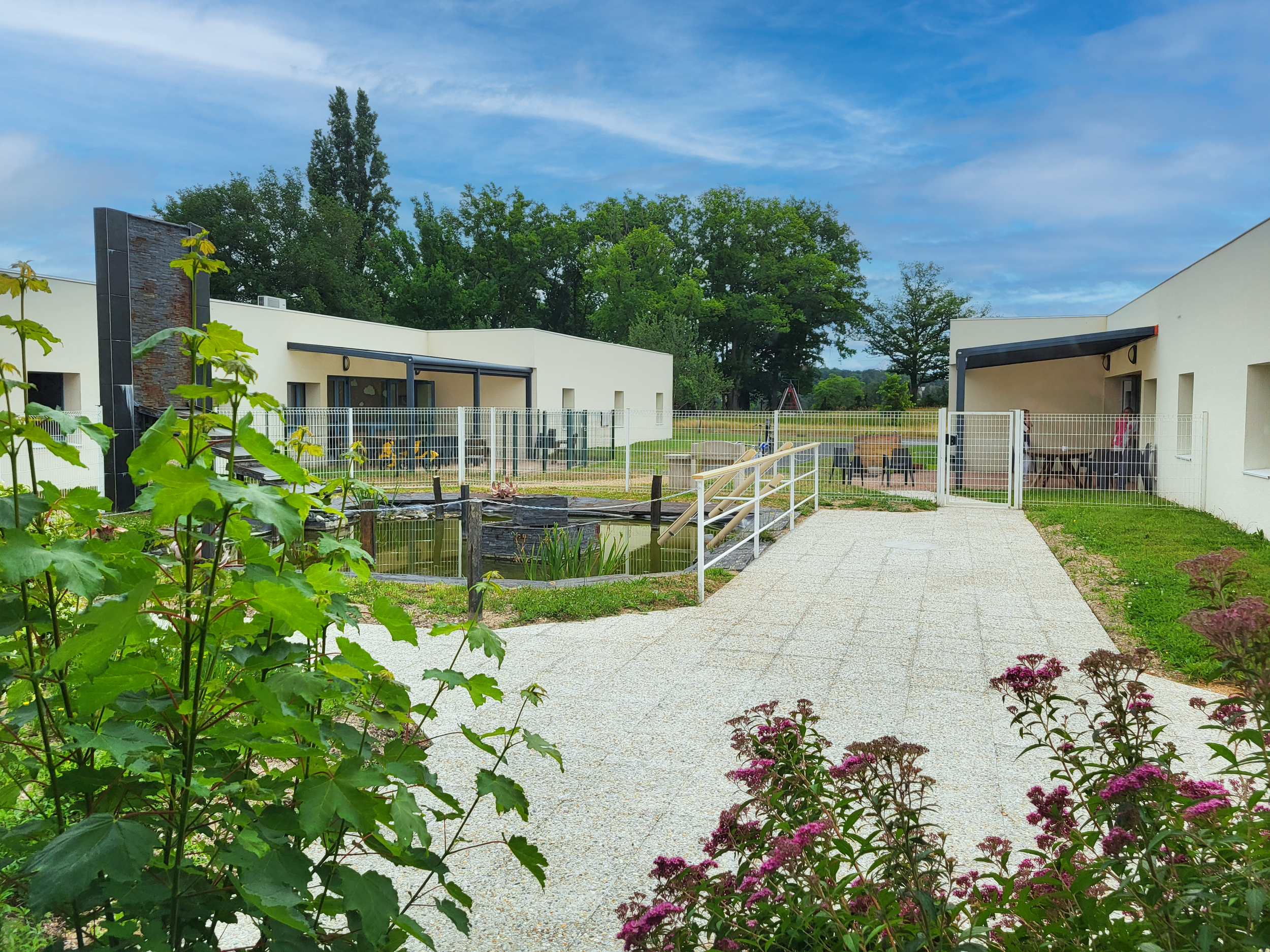 Centre d'accueil animalier avec bâtiments modernes, un enclos avec une petite piscine pour animaux, un chemin pavé, et des plantes en fleurs et en feuilles devant. Arbres verts et ciel bleu en arrière-plan.