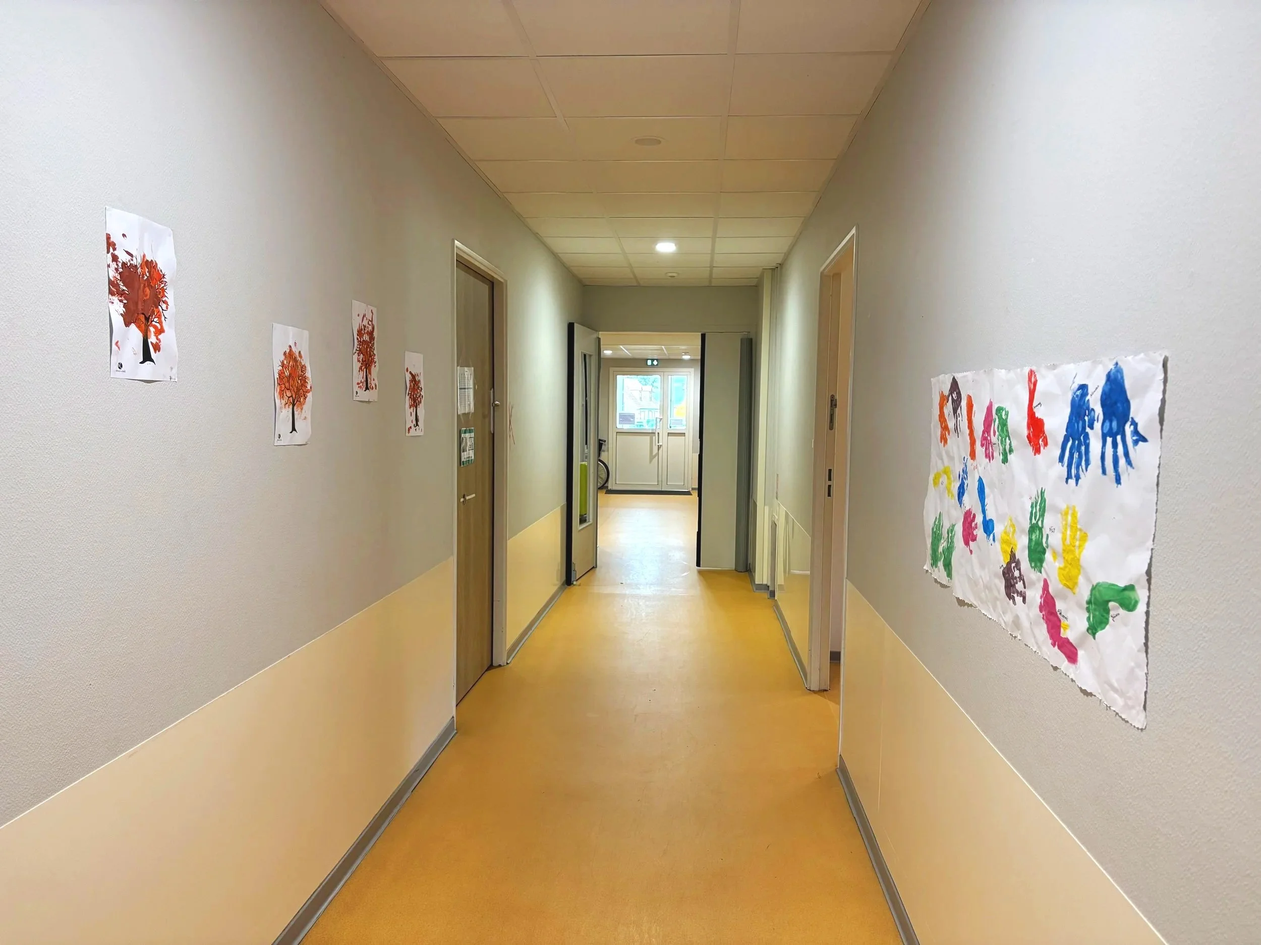 Couloir d'une école ou d'une garderie avec des dessins d'arbres en feuilles d'automne colorées sur le mur gauche et des empreintes de mains colorées sur le mur droit, avec une porte vitrée à la fin.