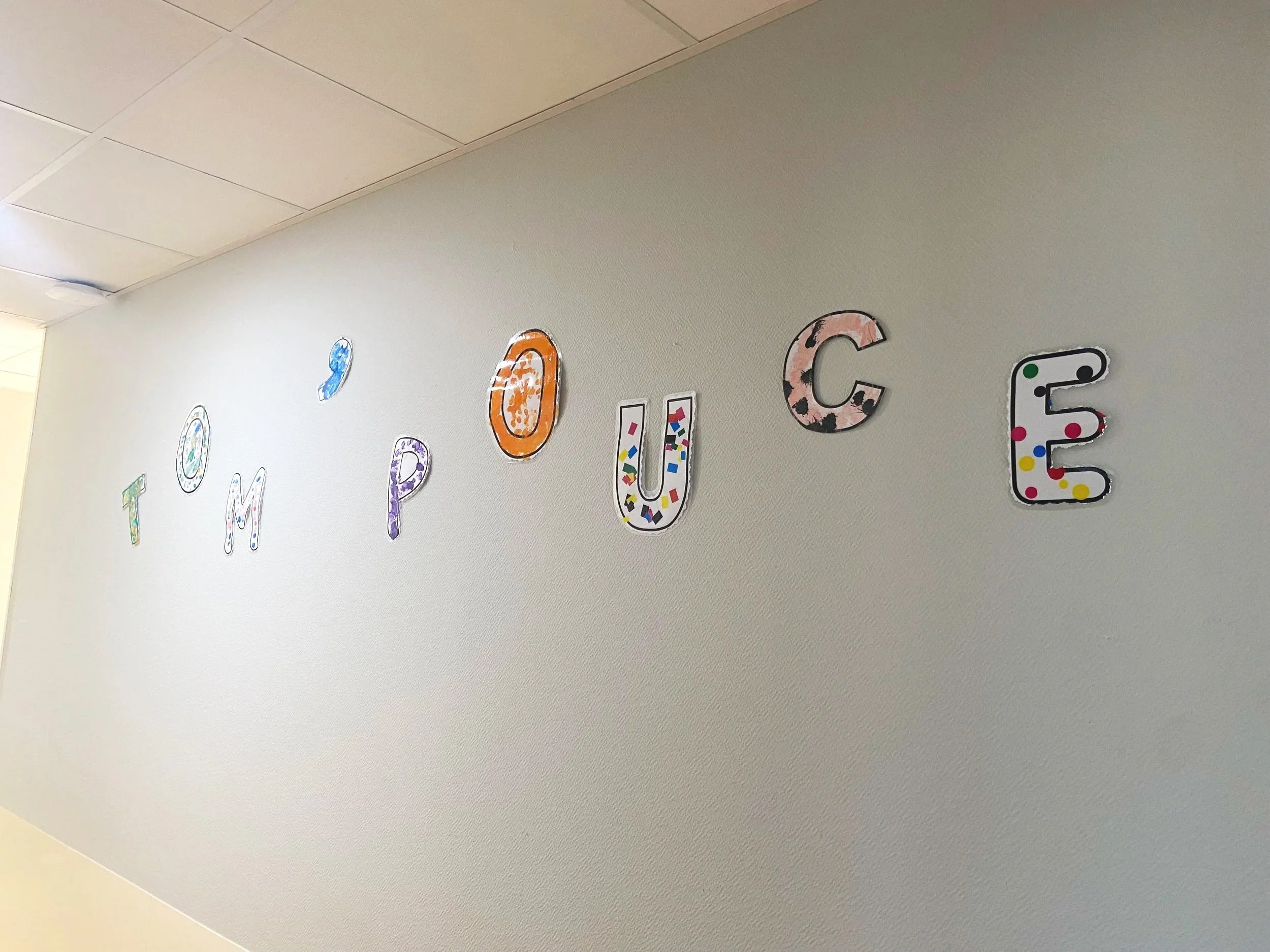 Les lettres formant le mot 'TOUMOUCÉE' décorées de diverses couleurs et motifs, accrochées sur un mur blanc.