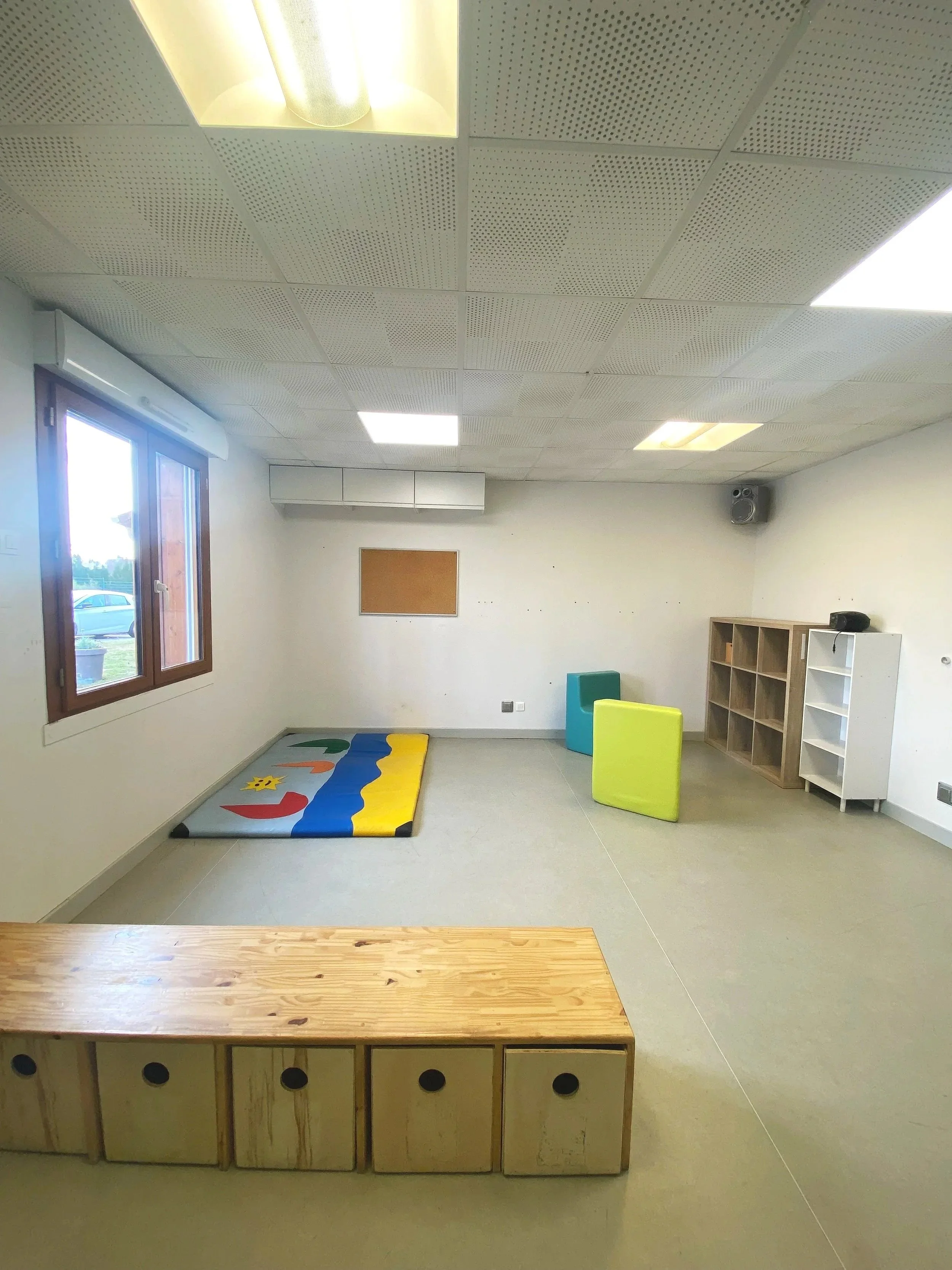 Une salle de jeux pour enfants avec un tapis coloré, des blocs en mousse, une étagère en bois et une fenêtre laissant entrer de la lumière naturelle.