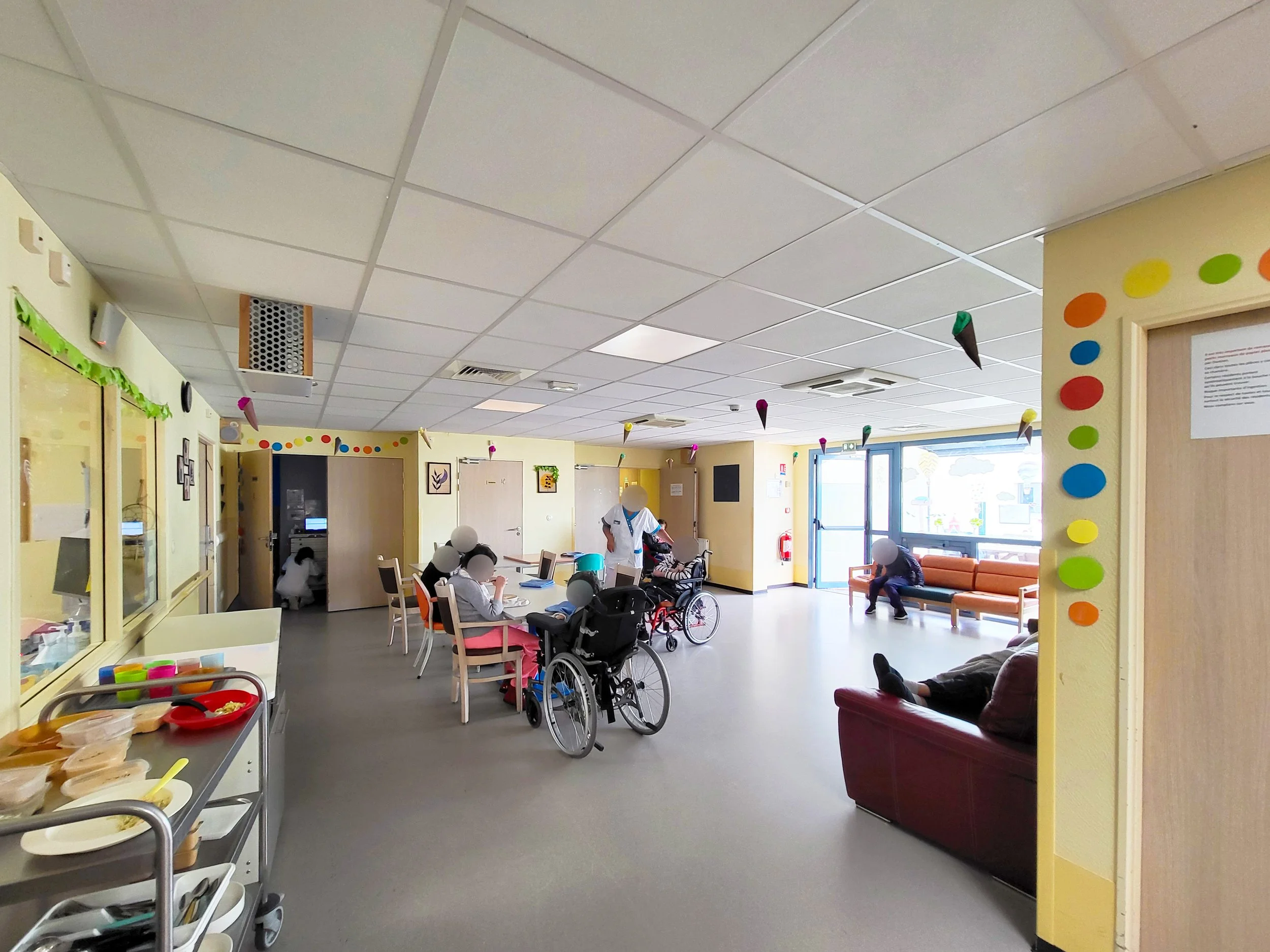 Salle de séjour dans un établissement de soins avec des personnes en fauteuil roulant et des chaises, décorée avec des cerises en papier suspendues.