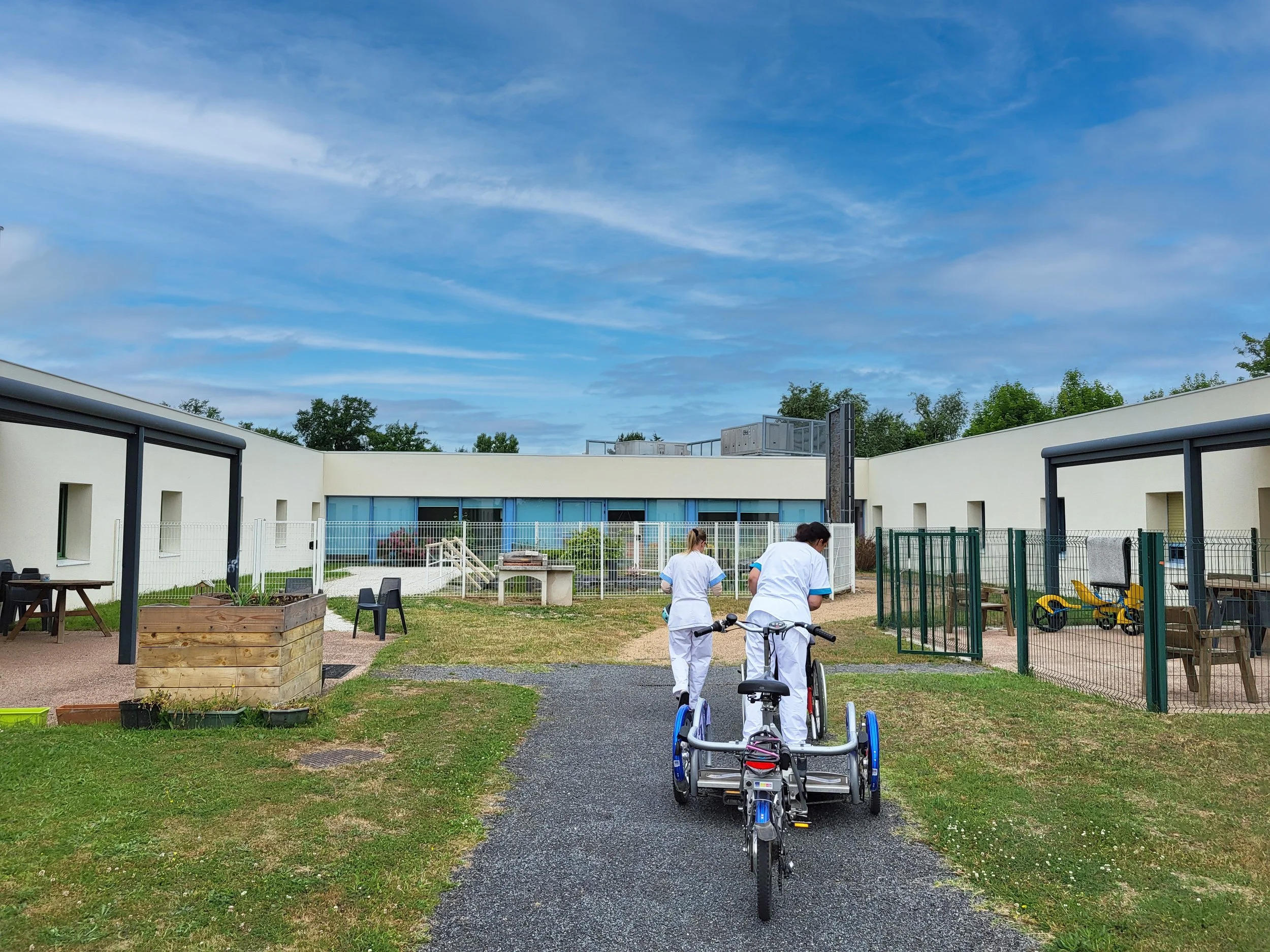 Deux professionnels en uniformes médicaux poussant un vélo adapté pour les personnes à mobilité réduite dans une cour extérieure en milieu hospitalier, entourée de bâtiments modernes blancs et bleus, avec un ciel bleu et quelques arbres en arrière-pl