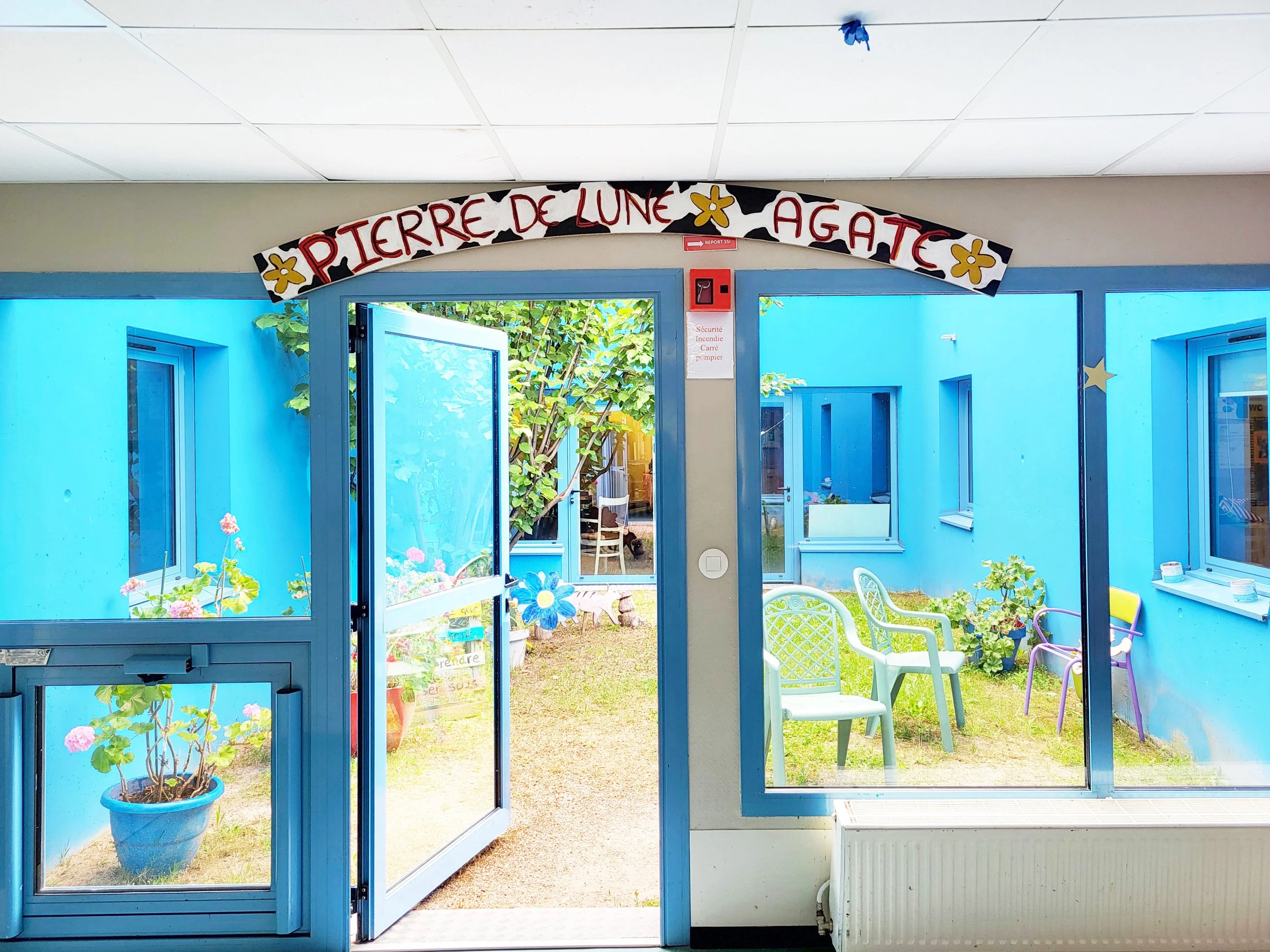 Cour intérieure avec chaise, plantes en pot, murs bleus, porte ouverte sur un jardin avec arbres.