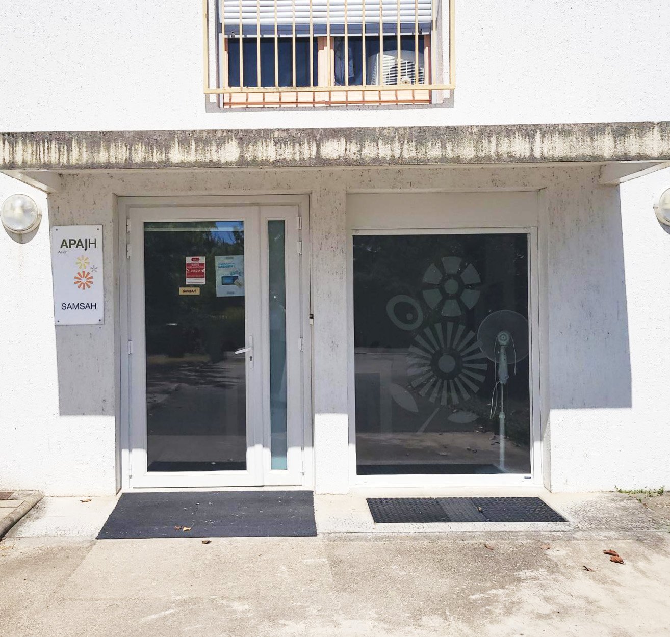 Façade d'un bâtiment blanc avec une porte en verre et une grande fenêtre, une affiche à gauche avec le texte 'APA JH' et 'SAMSAH', ainsi que des fleurs stylisées.