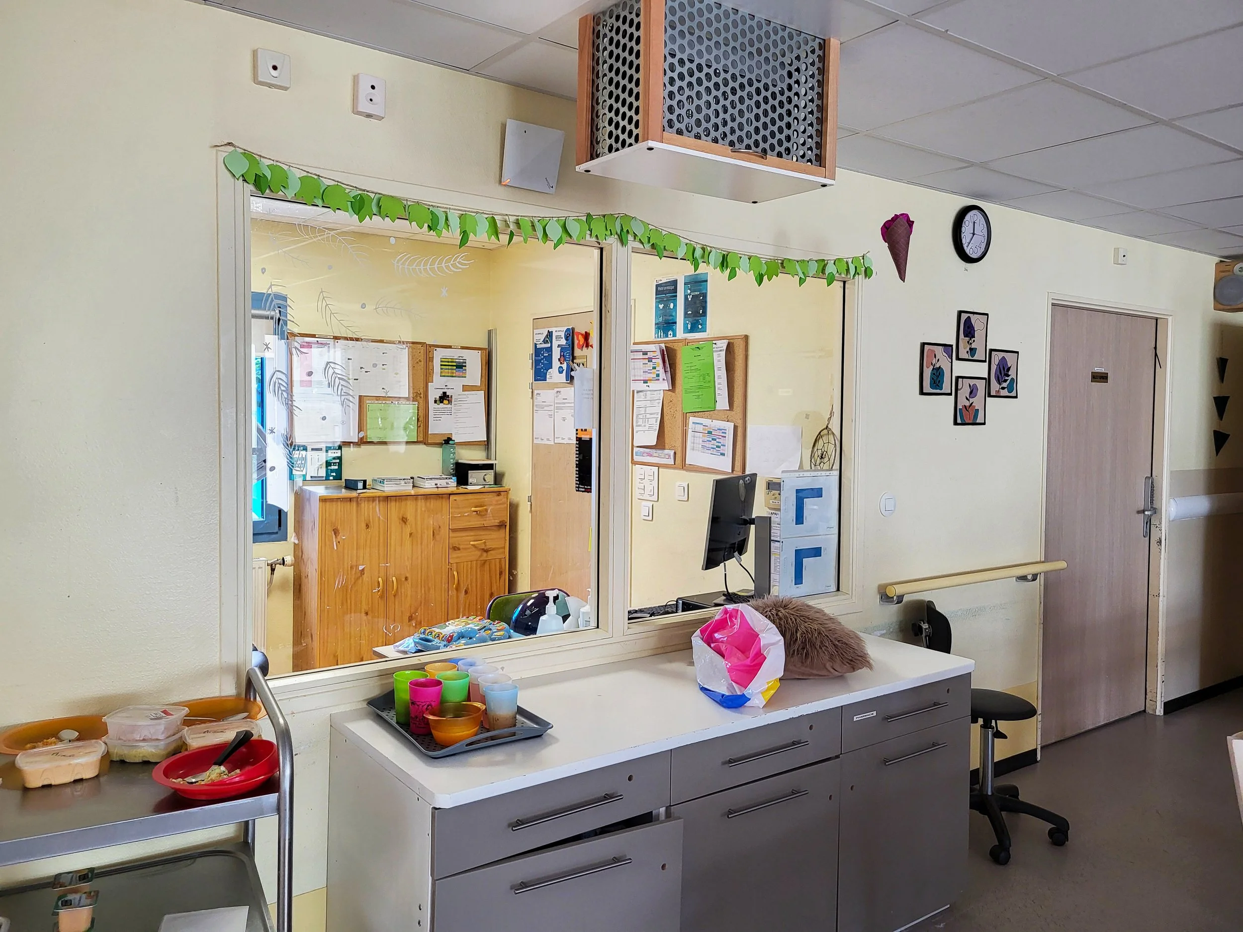 Une pièce d'un établissement scolaire ou de garde d'enfants avec un comptoir, des fournitures, un ordinateur et des décorations colorées. Vue à travers une fenêtre ou un panneau en verre.
