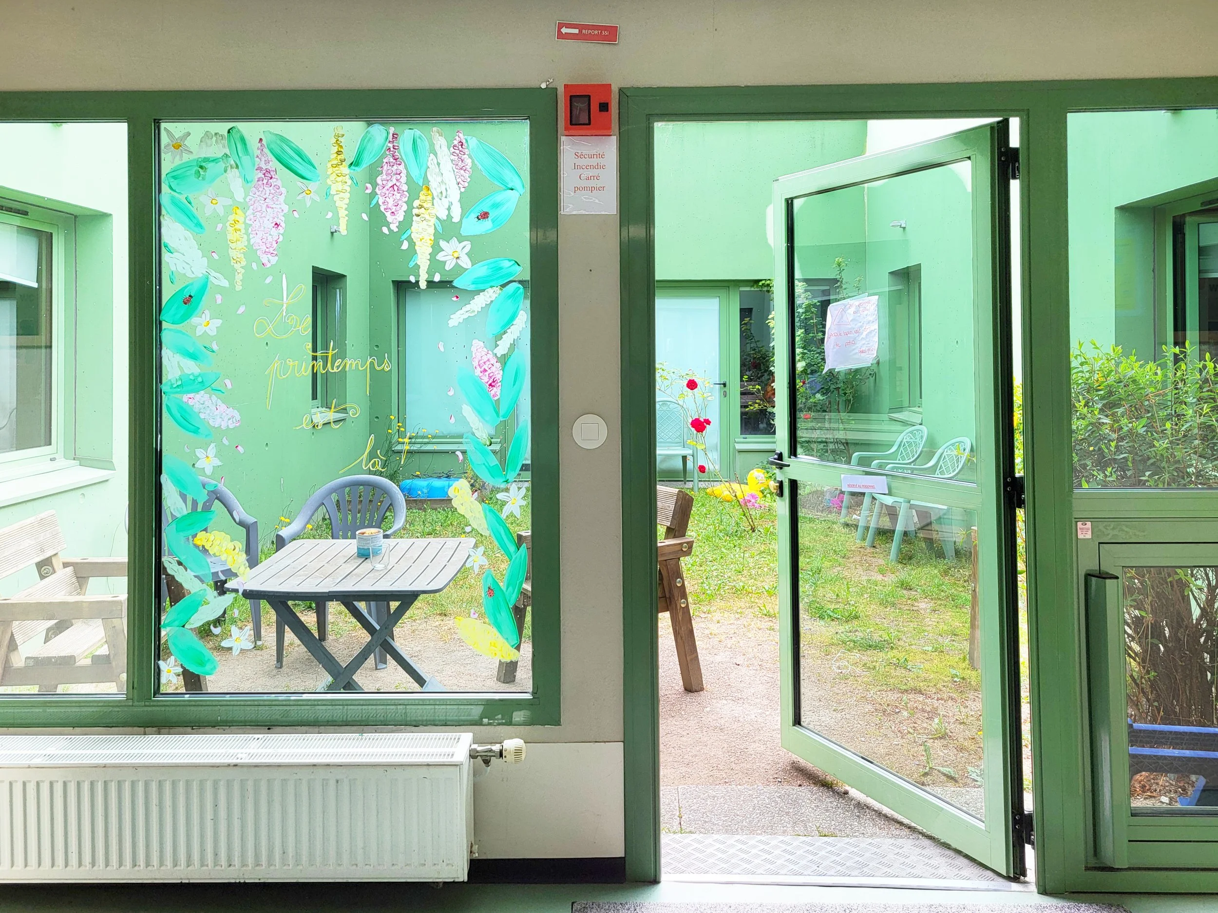 Vue depuis une fenêtre d'une terrasse avec mobilier en plastique, plantes et fleurs dans un jardin. Colombe de peinture, décoration florale et texte 'Le printemps est là' en lettres dorées sur la fenêtre