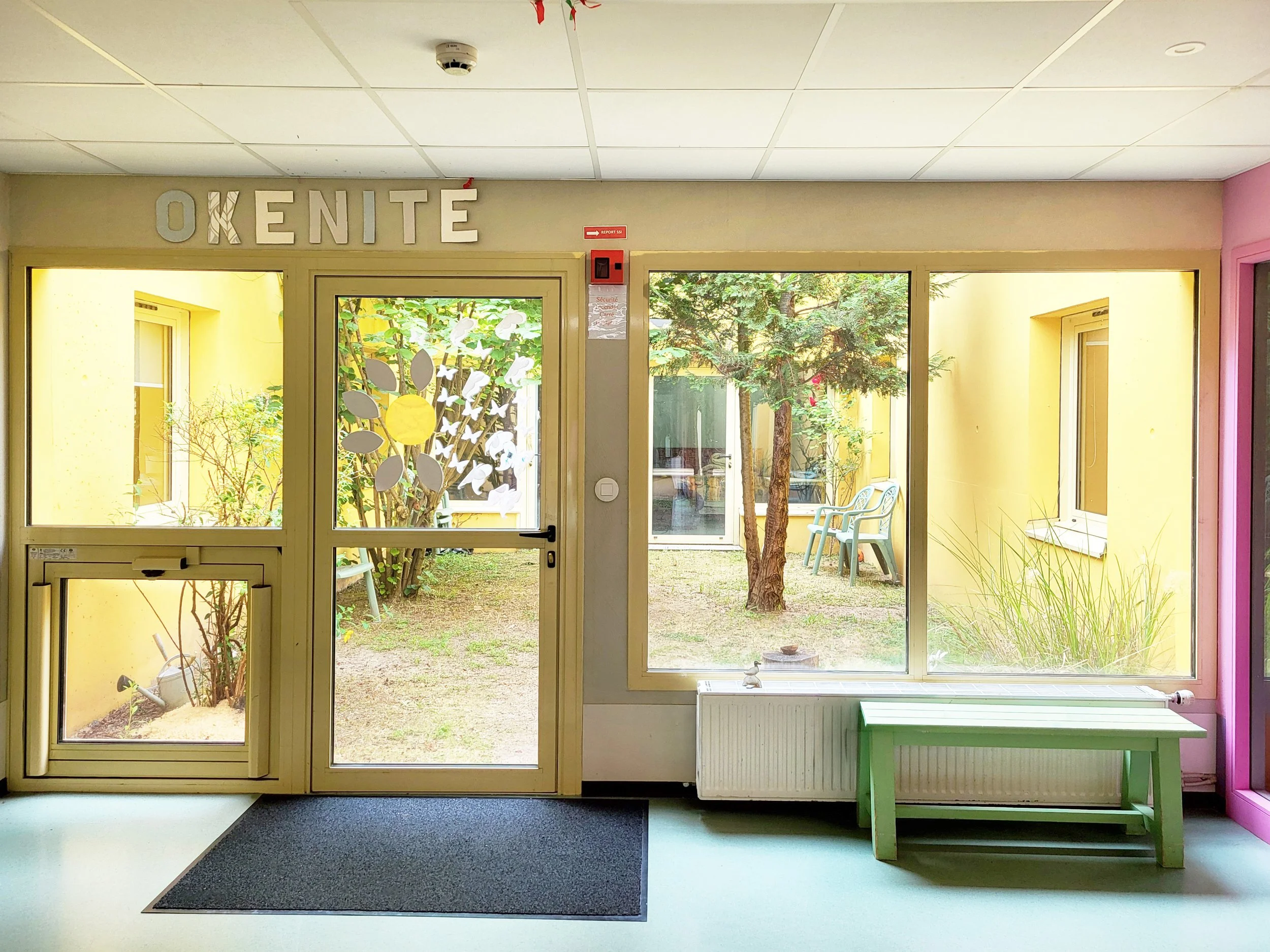 Vue intérieure d'une entrée de maison ou d'une garderie avec une porte en verre, vue sur un jardin avec arbres et plantes, murs jaunes, et un banc vert à côté d'une cheminée radiateur.