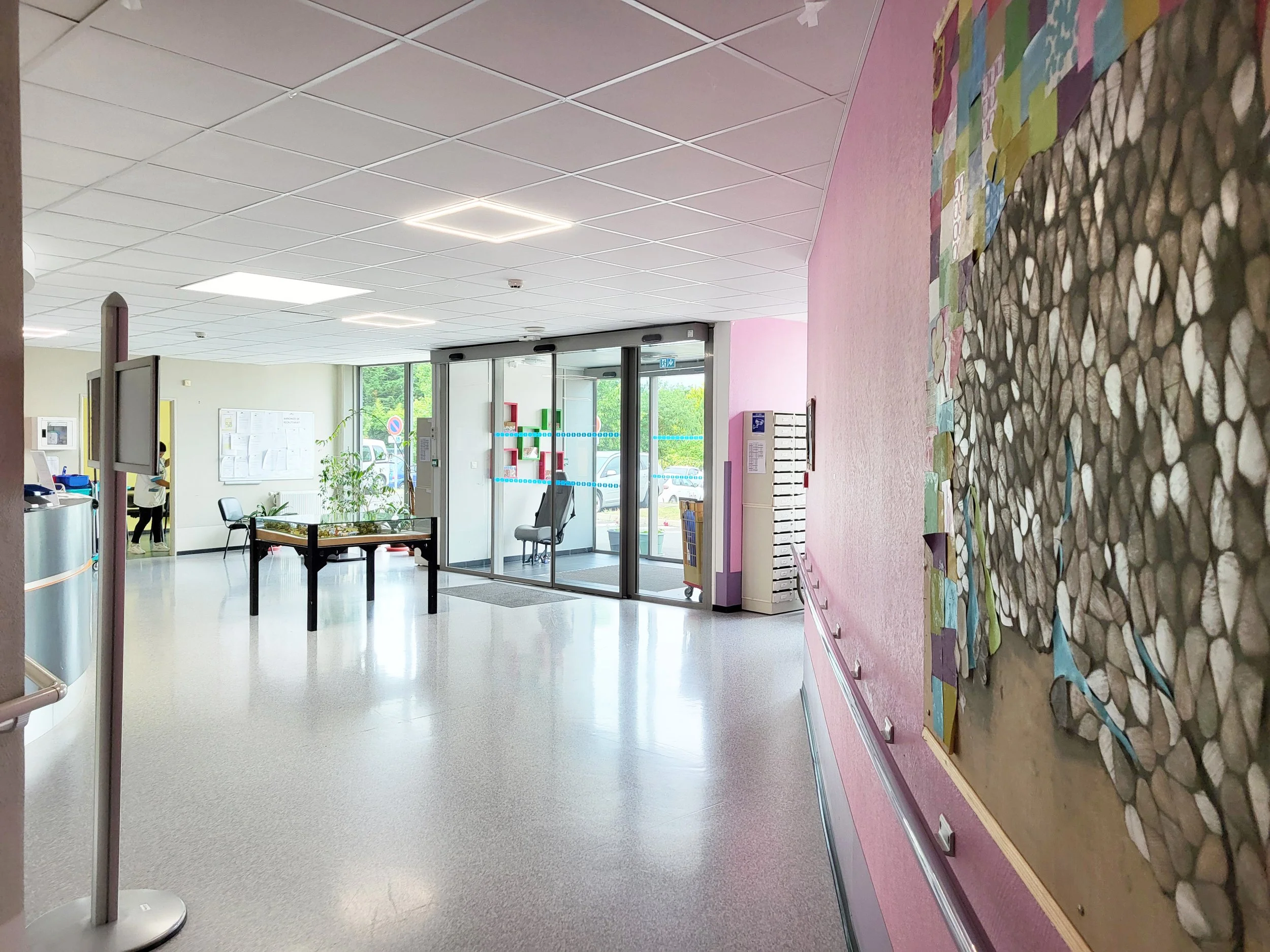 Hall d'accueil lumineux avec des murs aux couleurs pastel, une grande affiche d'information, un tableau d'affichage, une plante verte près de la porte d'entrée en verre, deux chaises, un comptoir d'accueil, et une œuvre d'art en mosaïque sur le mur.