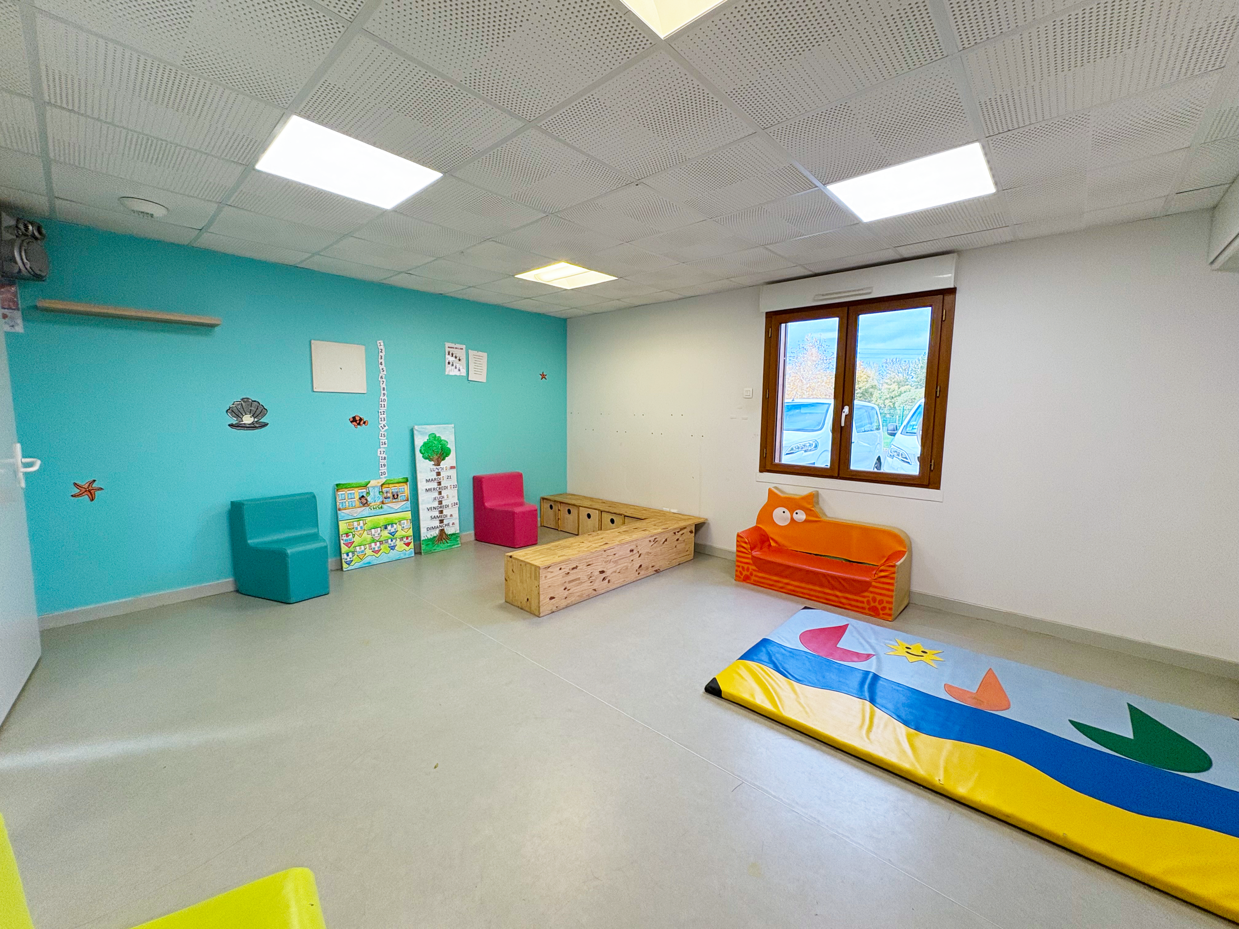 Salle de jeux pour enfants avec des bancs colorés, un tapis de jeu avec des formes, et des décorations marines sur un mur bleu.