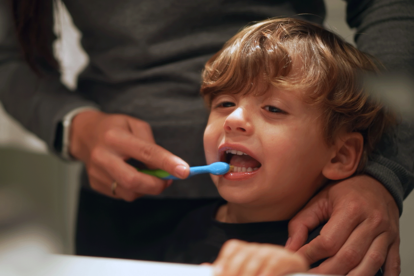 Sad little boy having his teeth brushed from his parent.