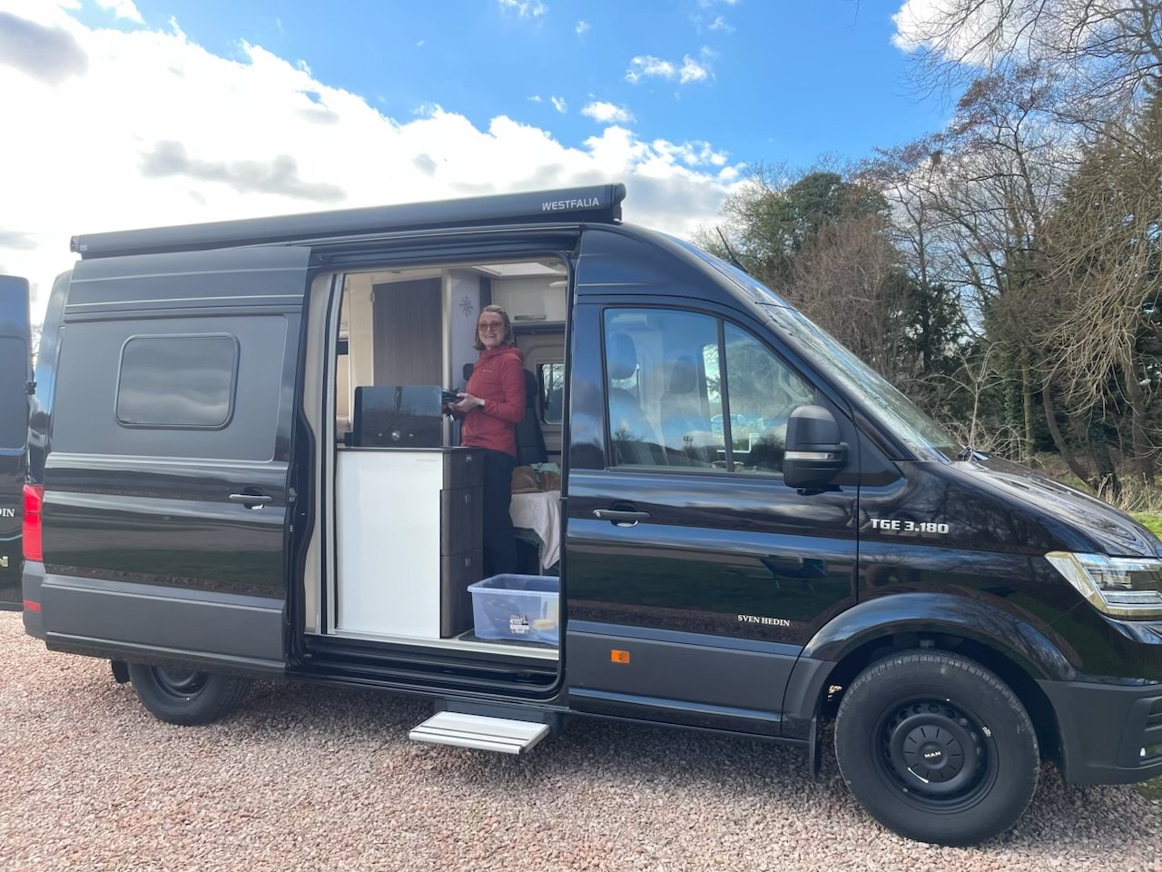 Black camper van parked outdoors with sliding door open, showing interior and a person standing inside near a kitchen area.