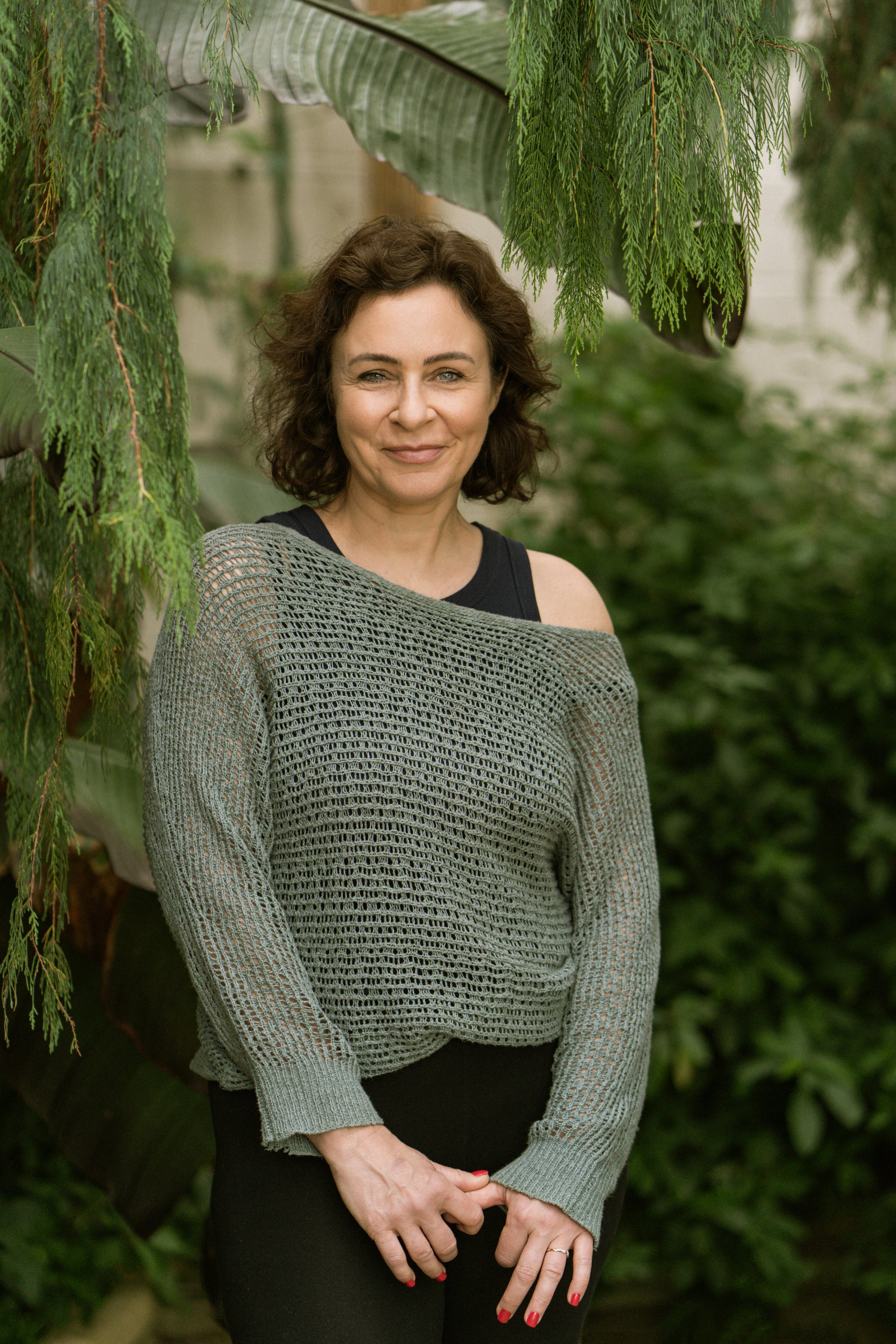 A woman with short curly brown hair standing outdoors among green plants, wearing a black sleeveless top and a gray open-weave cardigan, smiling at the camera.