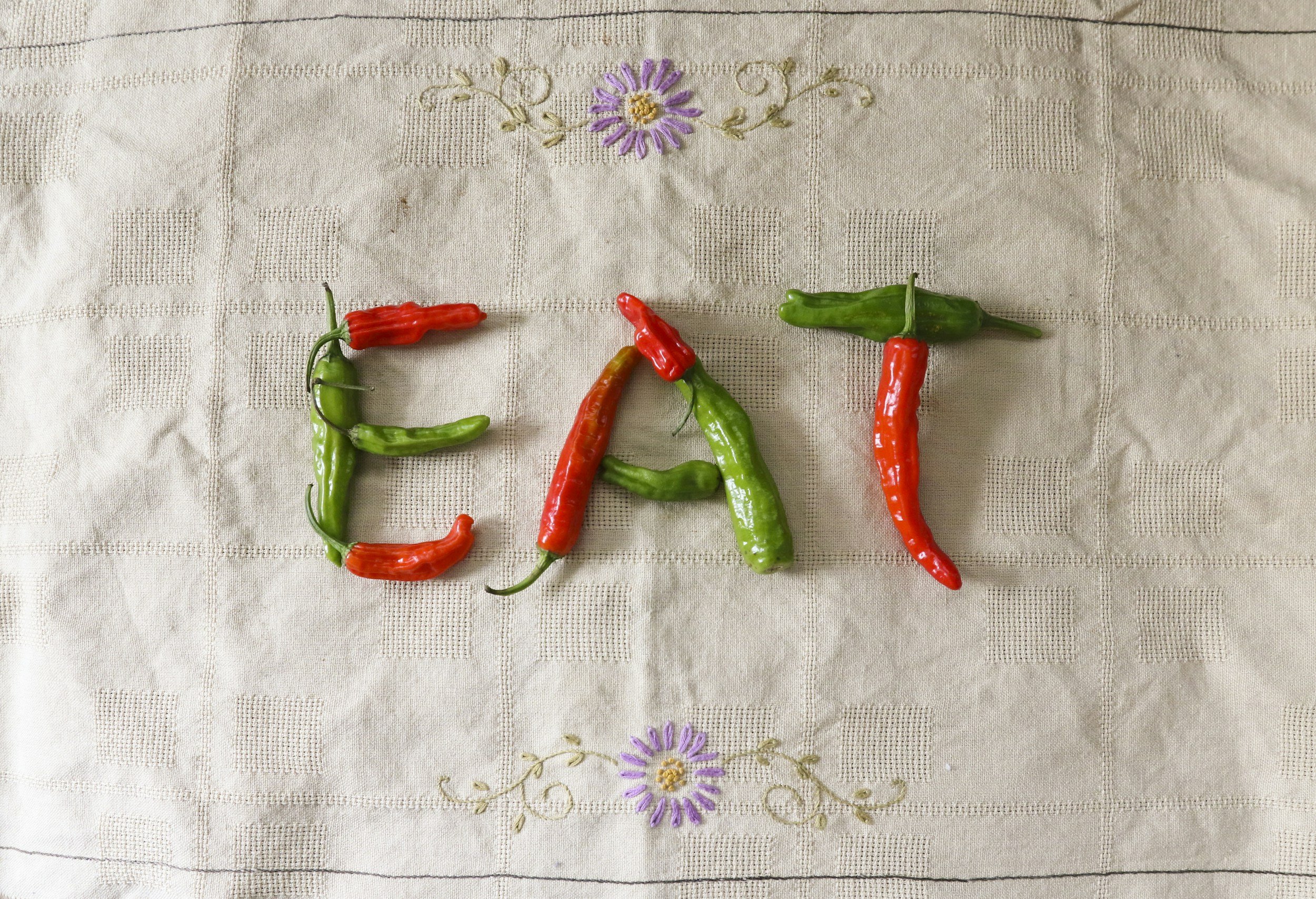 Chili peppers arranged on a tablecloth spelling out the word 'EAT' with decorative purple flowers above and below.