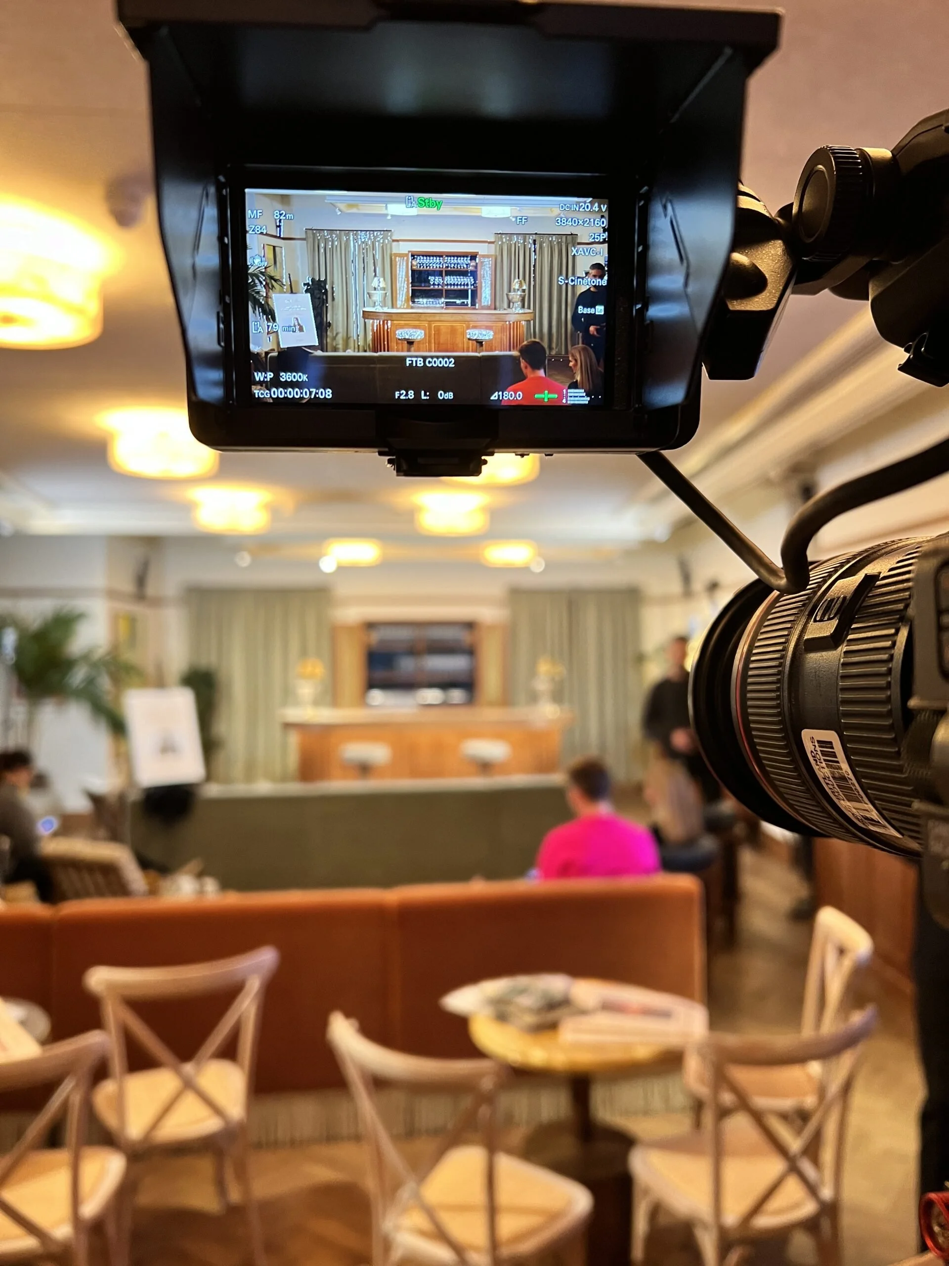 A camera on a tripod pointing at a stage with a bar counter in a cozy, well-lit room. The room has rows of chairs and tables, and a few people are seated, facing the stage.
