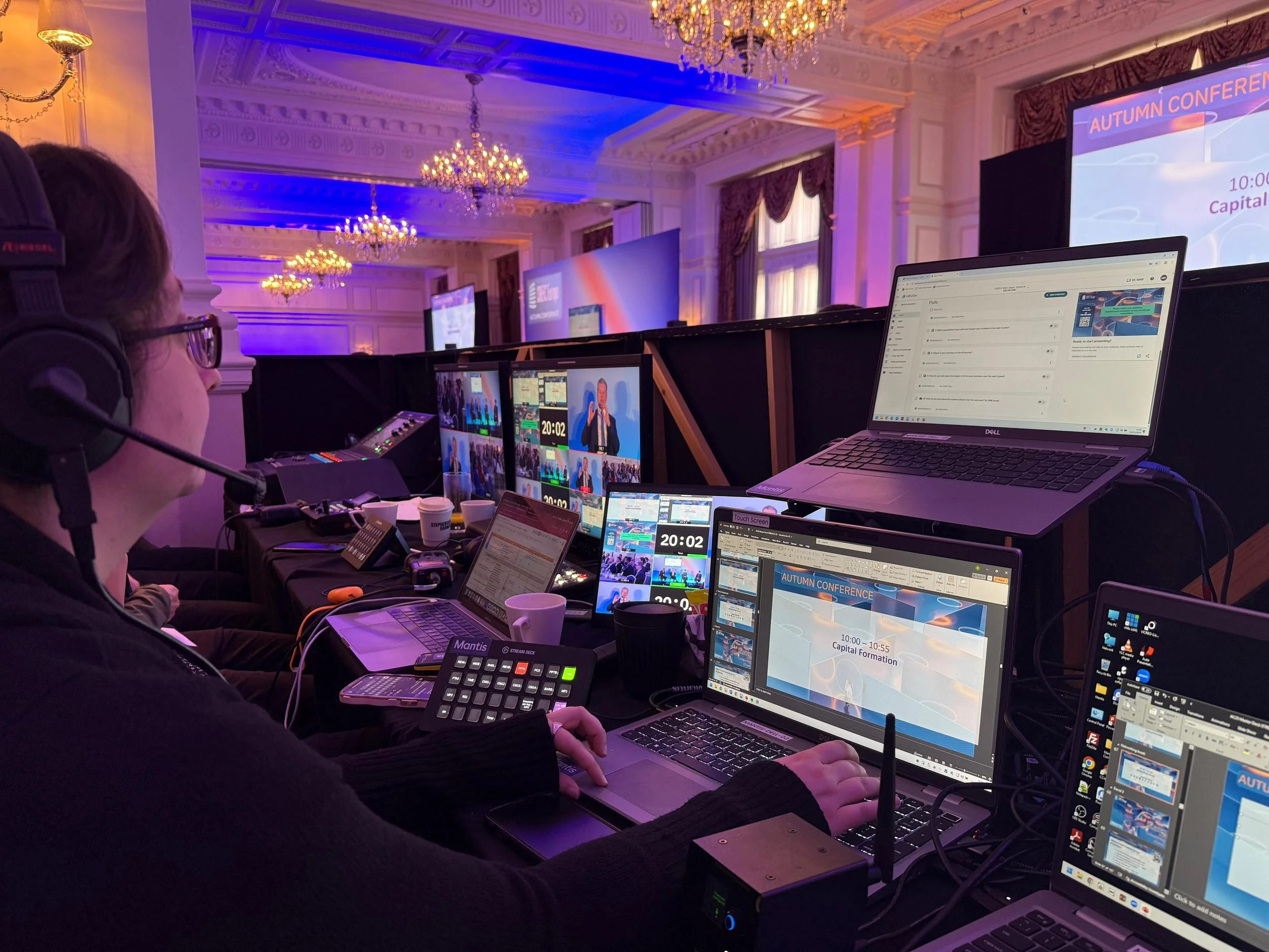 A person wearing headphones is working at a control station with multiple laptops, monitors, and equipment during a conference event in a large ornate room with chandeliers and colorful lighting.