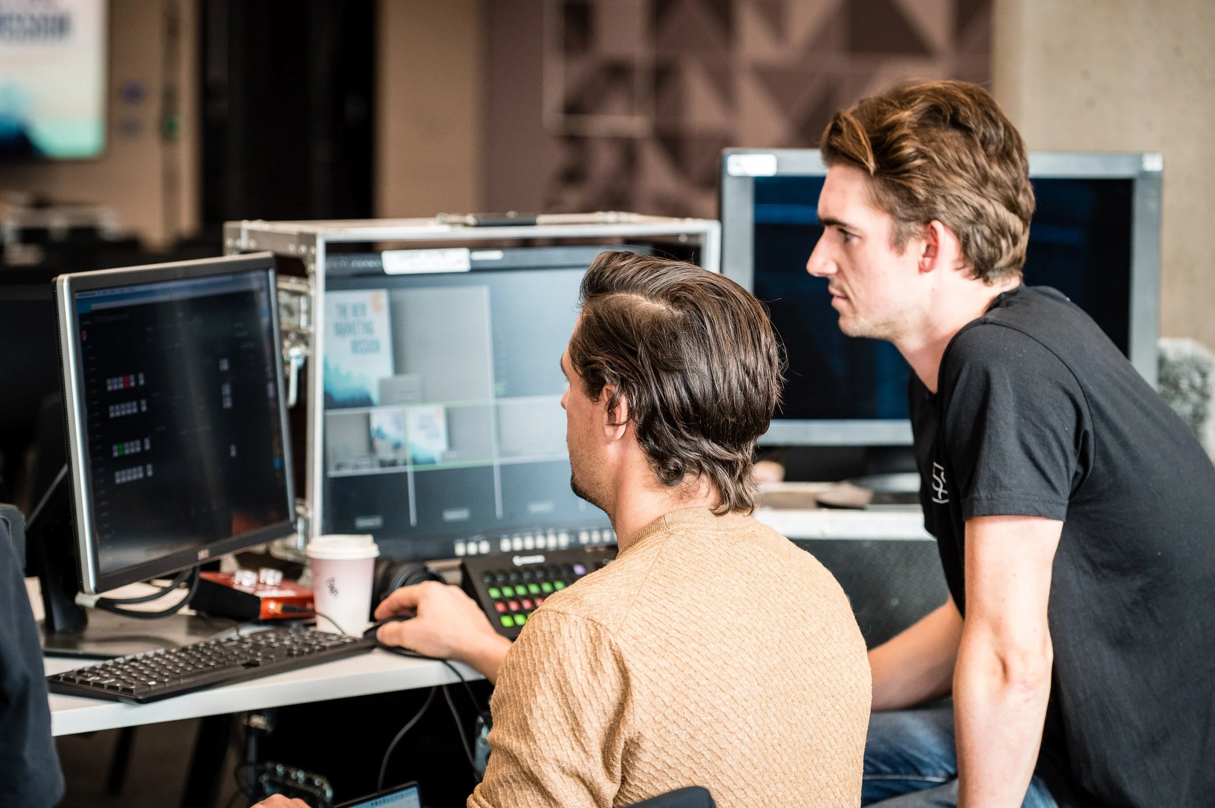 Two engineers working on computers in a tech environment, with multiple monitors, one with a video editing software, and a coffee cup on the desk.