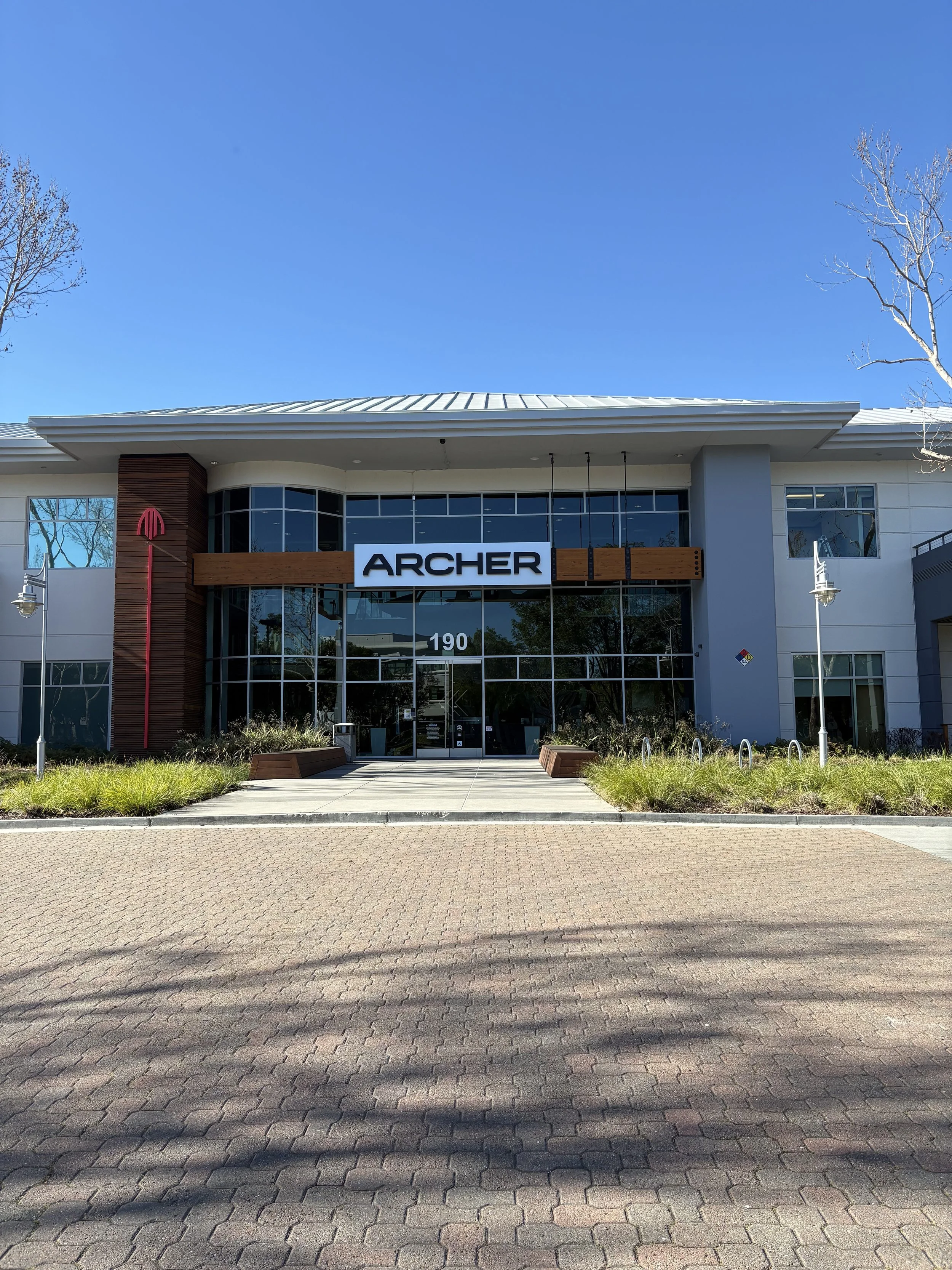 Front view of a modern commercial building with large glass windows, a sign that reads 'ARCHER,' and surrounding landscaped area with grass and trees under a clear blue sky.