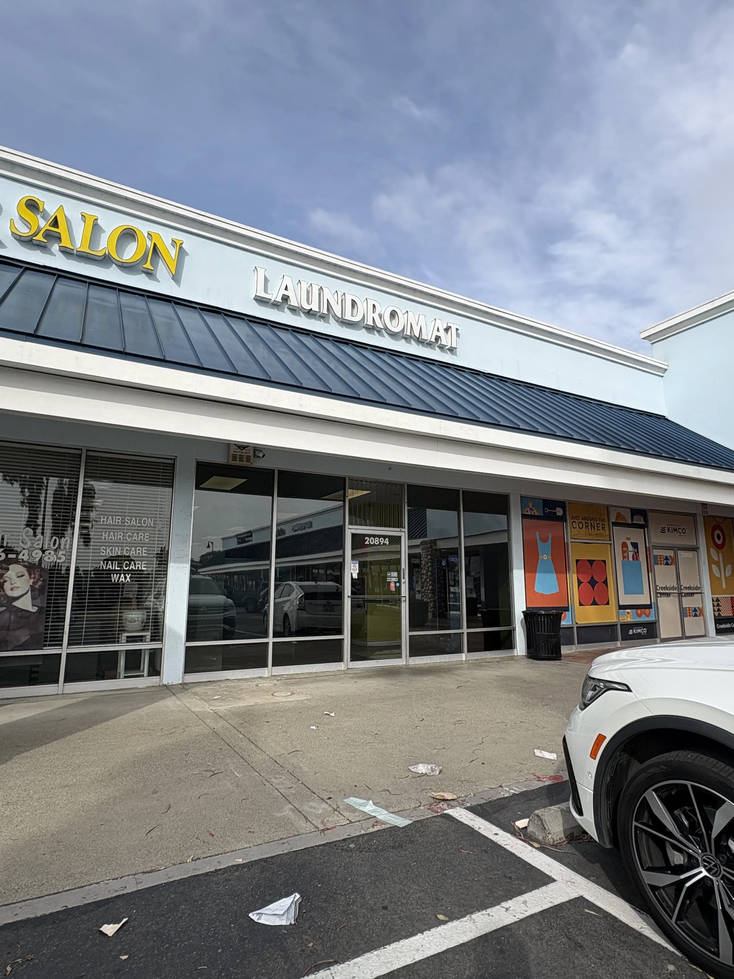 The exterior of a shopping plaza with a sign for a salon and laundromat, a glass storefront, and a parking lot with a white car, litter on the ground, and commercial posters.