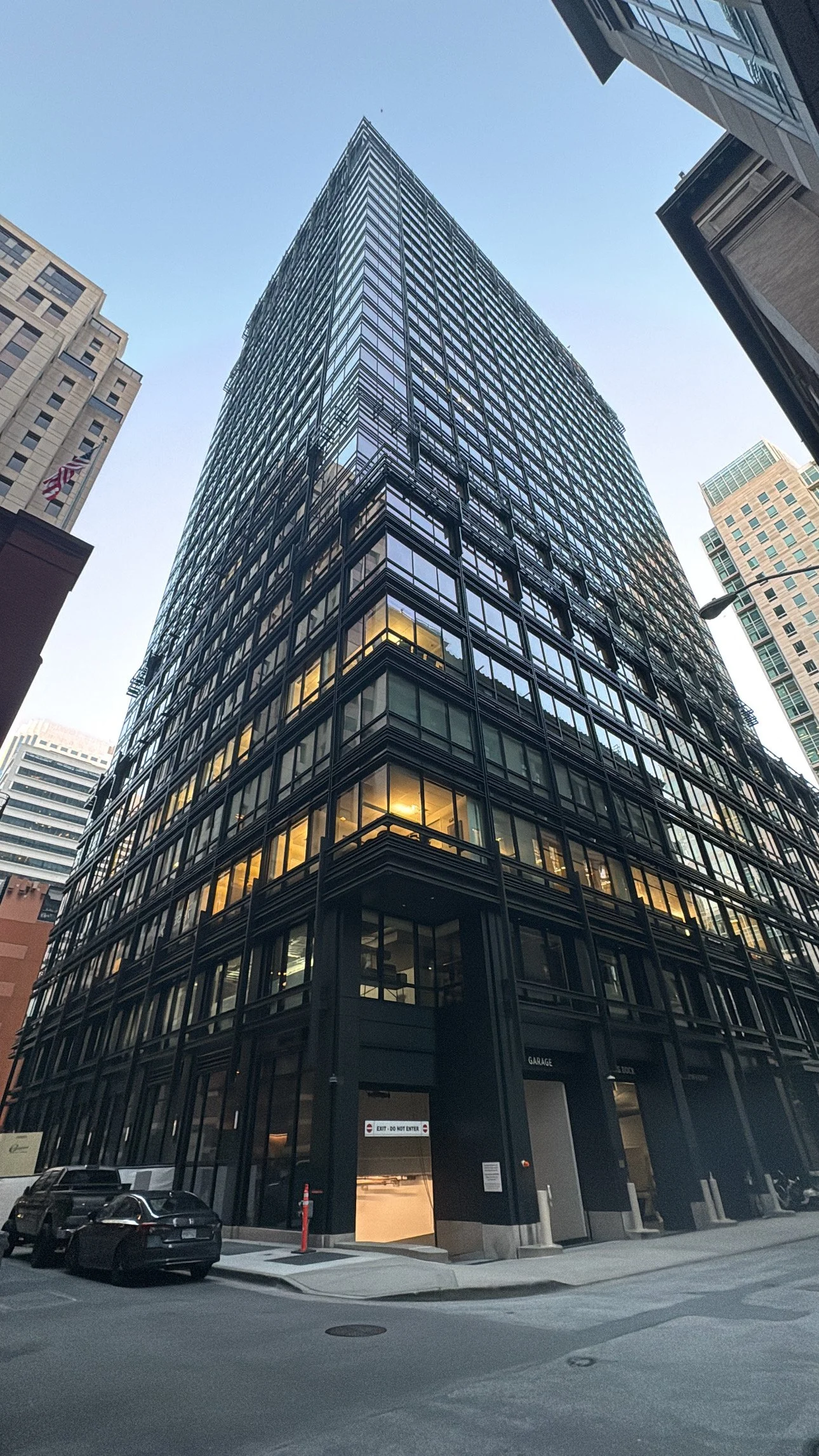 A tall black glass building viewed from street level, with illuminated office windows at the lower levels and neighboring buildings in the background. The sky is clear and blue.
