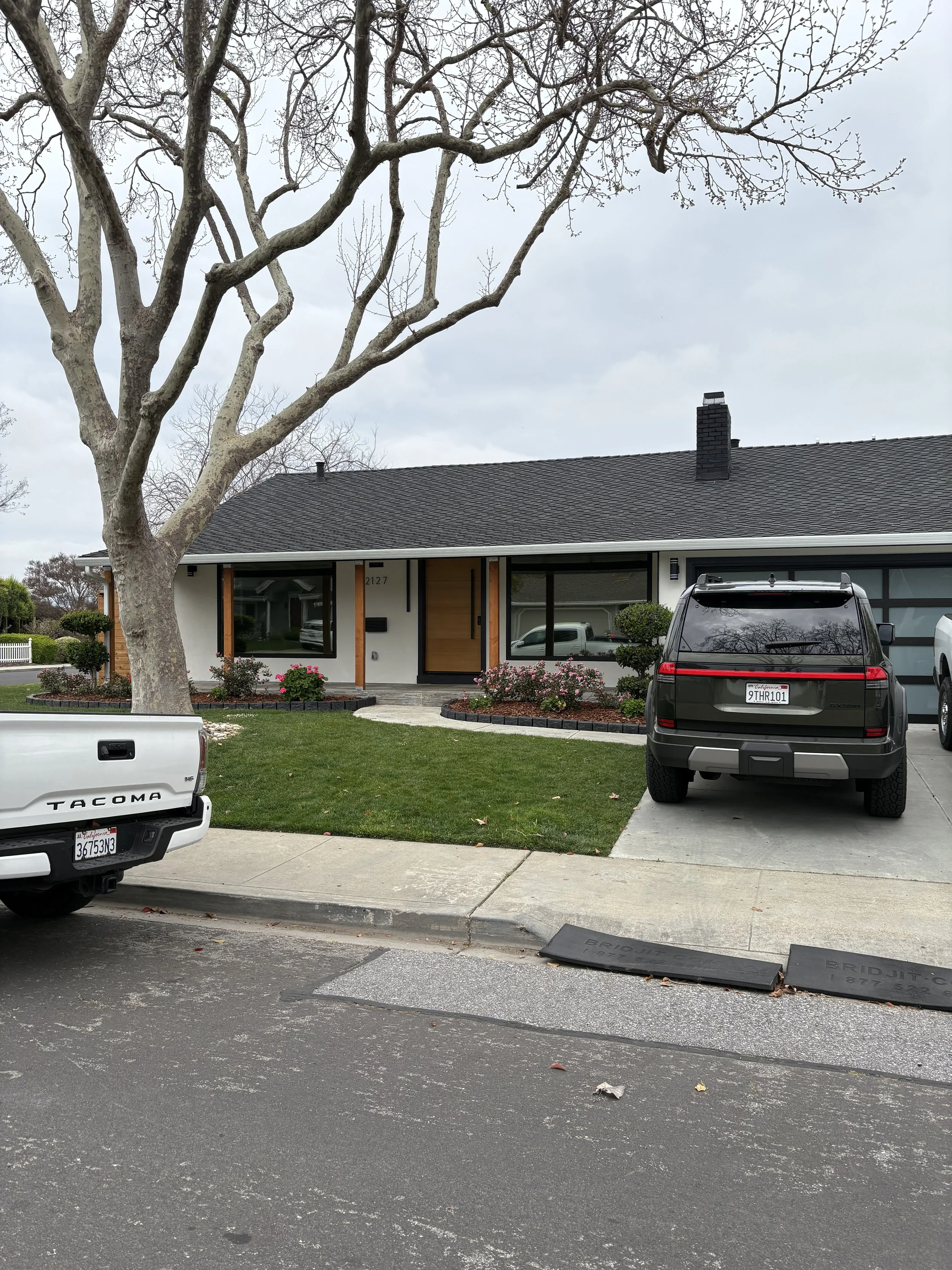 Front view of a modern house with cars parked in the driveway and a large tree in the front yard under a cloudy sky.