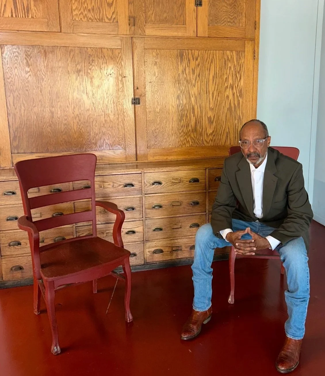 Bobby Henderson wearing glasses, a green blazer, white shirt, blue jeans, and brown boots sitting on chairs from the Omaha Stock Yard