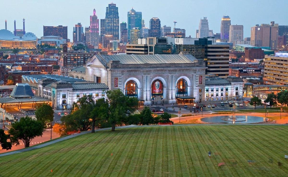 Nighttime city skyline with tall buildings and neon lights, overlooking a historic train station with classical architecture, trees, parking lot, and a grassy foreground in Kansas City, Missouri.
