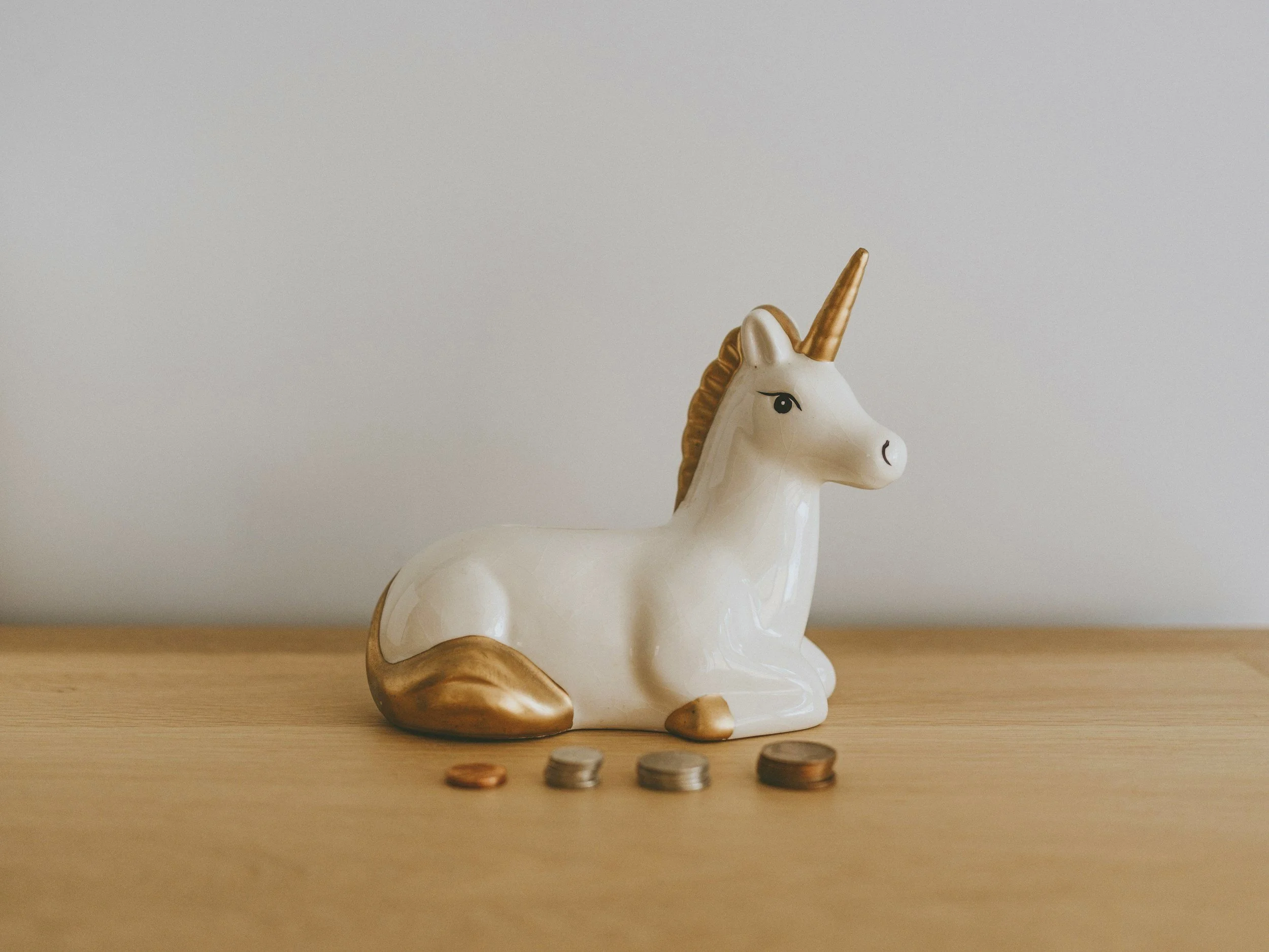 A ceramic unicorn piggy bank with a white body, gold horn, tail, and mane, sitting on a wooden surface with four coins in front of it.