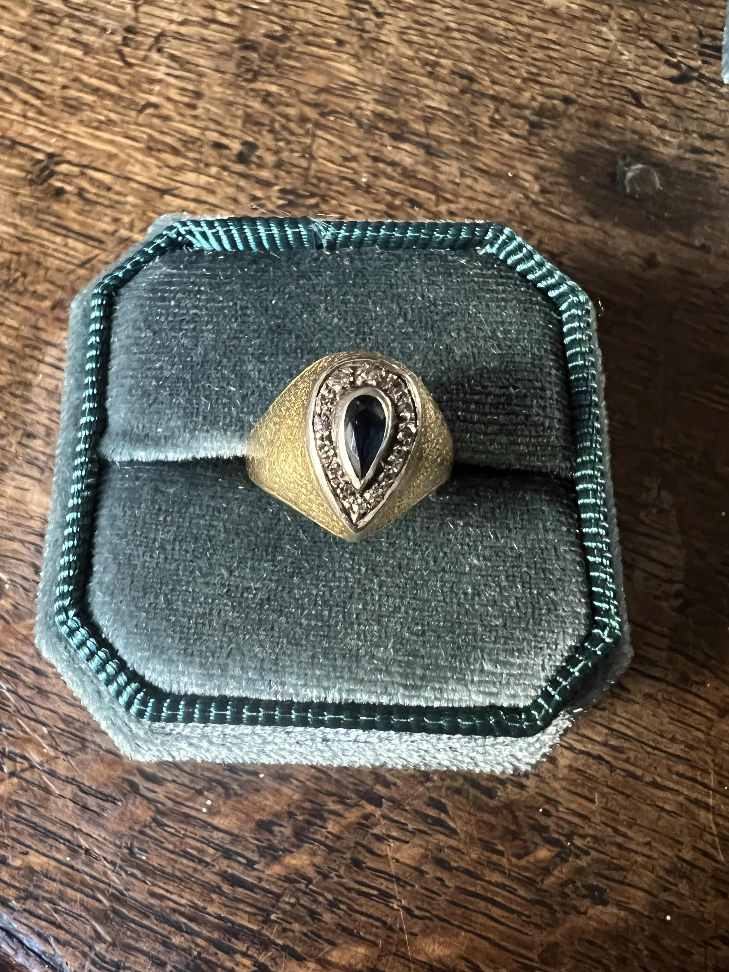 Une bague en or avec une pierre noire centrale entourée de petites pierres, dans un coffret en velours gris. 