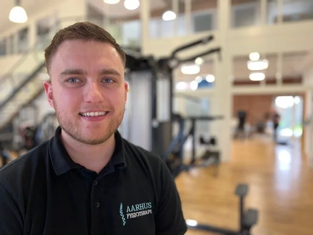 A young man with short brown hair and a beard, smiling at the camera, wearing a black polo shirt with 'AARHUS FYSIOTERAPI' logo, in a gym or rehabilitation center with exercise equipment in the background.