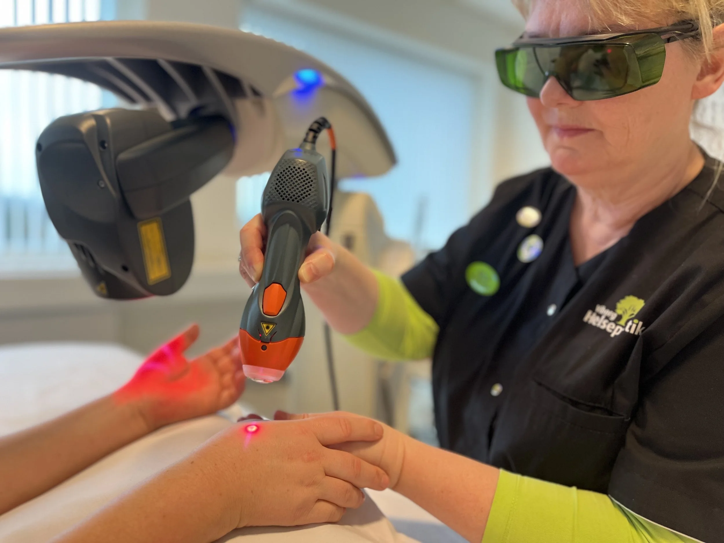 A healthcare professional performing laser therapy on a patient's hand in a clinical setting, both wearing protective glasses.