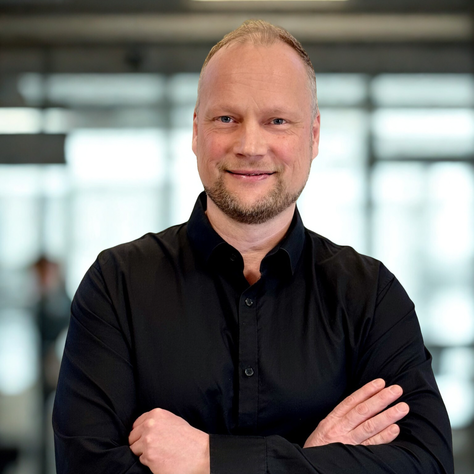 Mand i sort skjorte, mellemblondt hår og skæg, står med armene over kors og smiler, New Yorker glasrude baggrund ,Christian Bjørkman