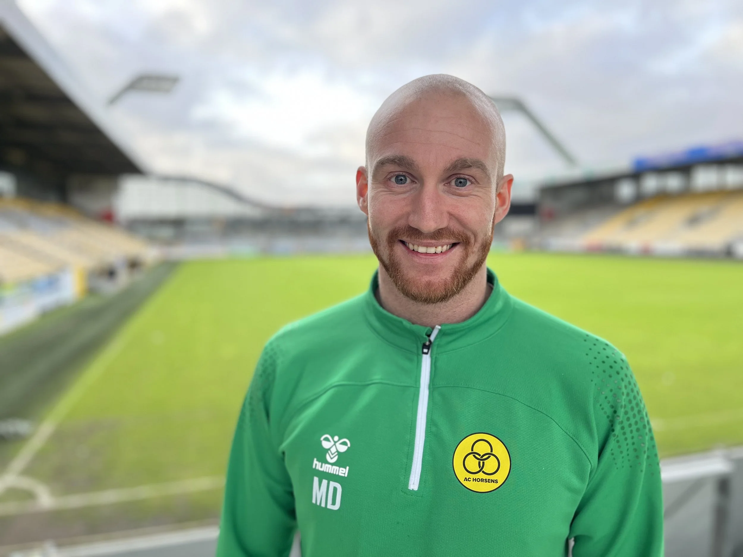 A man with short hair and a beard, smiling, wearing a green AC Horsens sports jacket with the club's logo on it, standing in a football stadium with empty stands and a green field in the background.