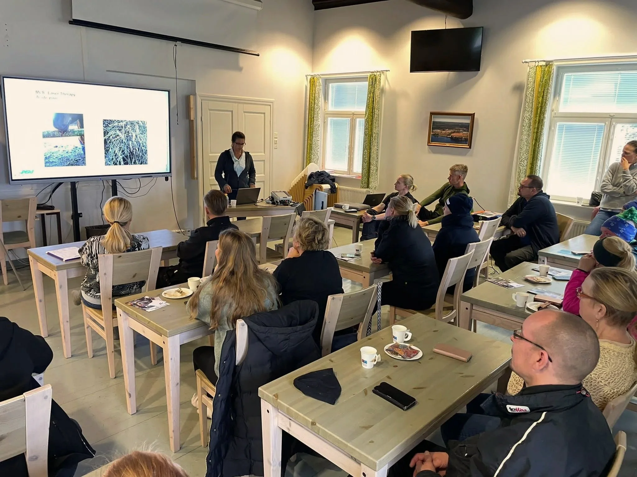 En flok voksne mennesker sidder og ser op på en skærm i et undervisningslokale. Der står en underviser en kvinde med en laptop foran sig. På bordene står der kappekopper, blokke og en mobiltelefon.