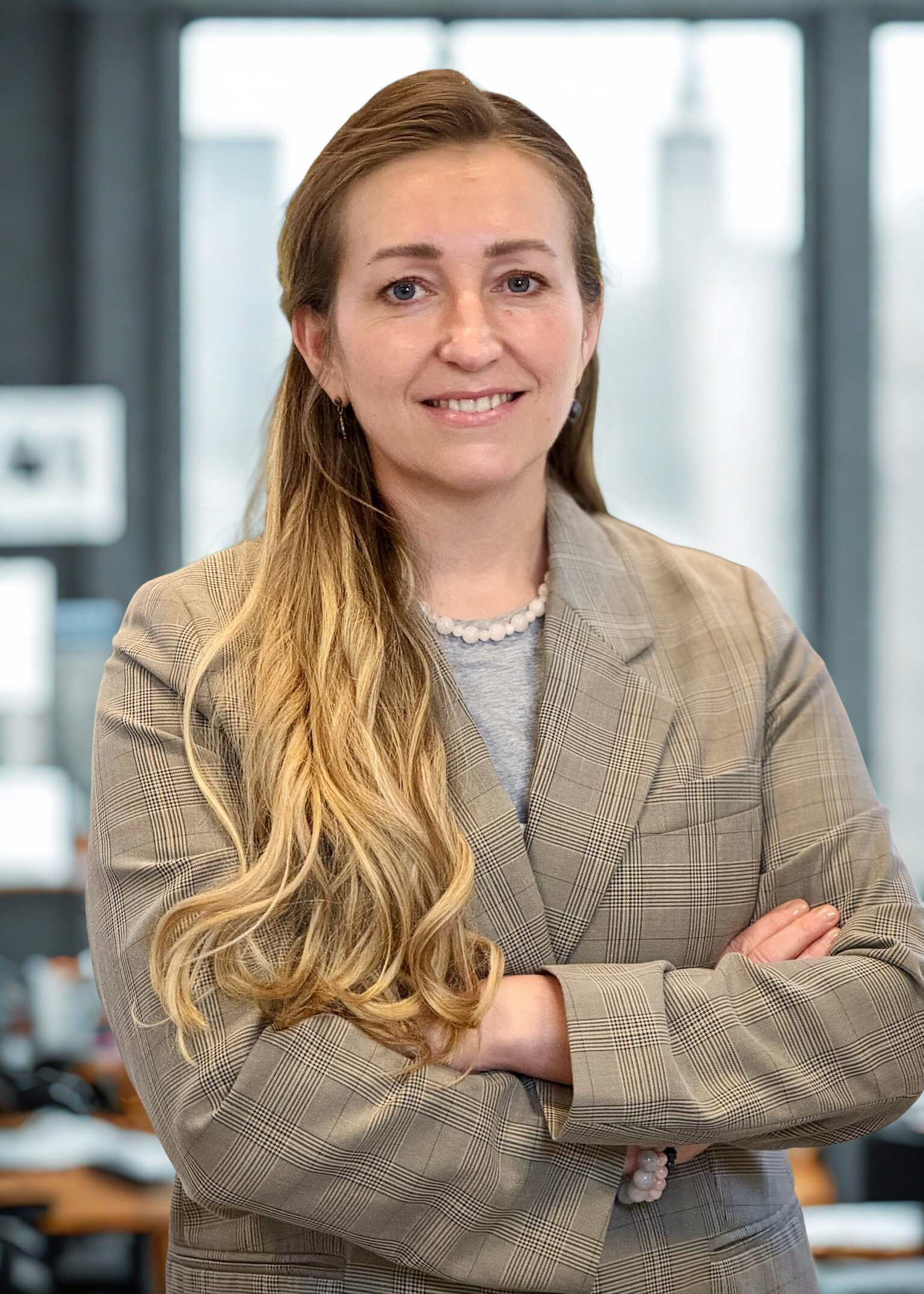 A woman with long blonde hair and wearing a blazer and necklace,  smiling in front of a New Yorker glas wall.