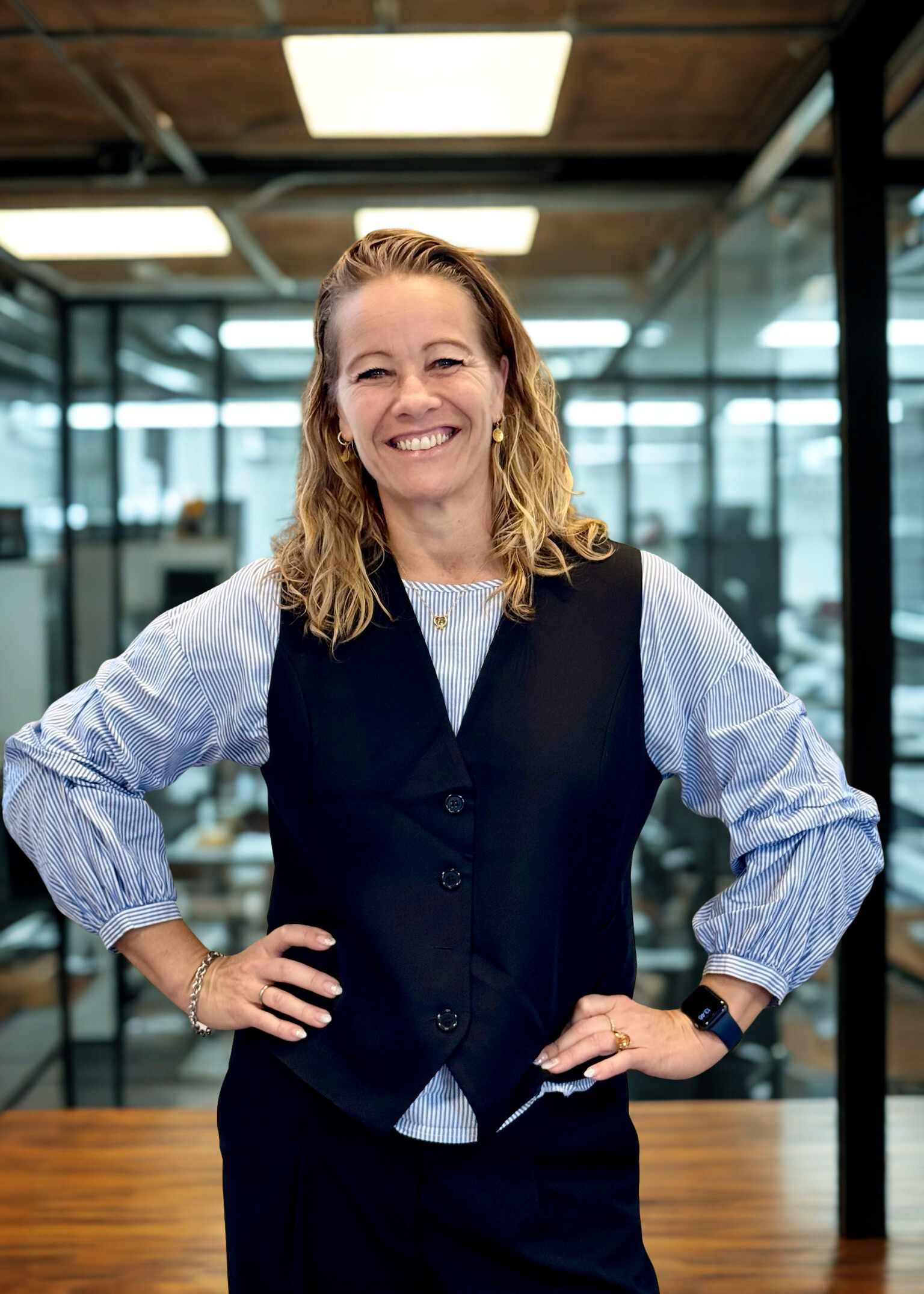 Kvinde med mellemlangt mellemblondt hår og stort smil, står med hænderne i taljen og har en stribet skjorte på og sort vest ud over, I baggrunden ses et kontormiljø og New Yorker glas ruder, Louise Reisz