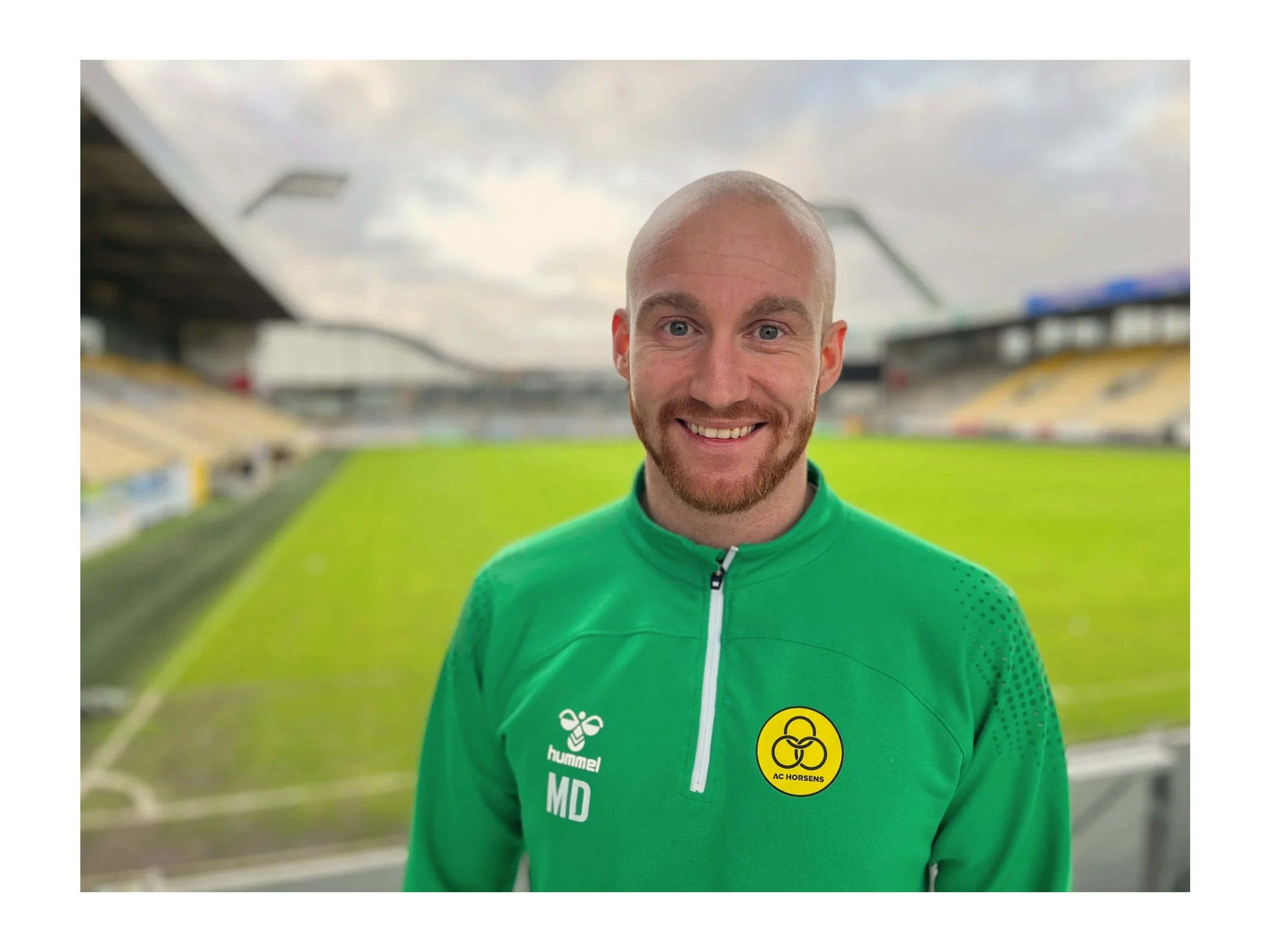 A man with short hair and a beard, smiling, wearing a green AC Horsens sports jacket with the club's logo on it, standing in a football stadium with empty stands and a green field in the background.