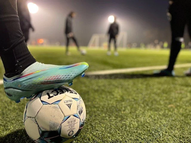 A soccer player balancing a soccer ball with their foot on a field at night. Several other players are in the background, and the field is lit with floodlights.
