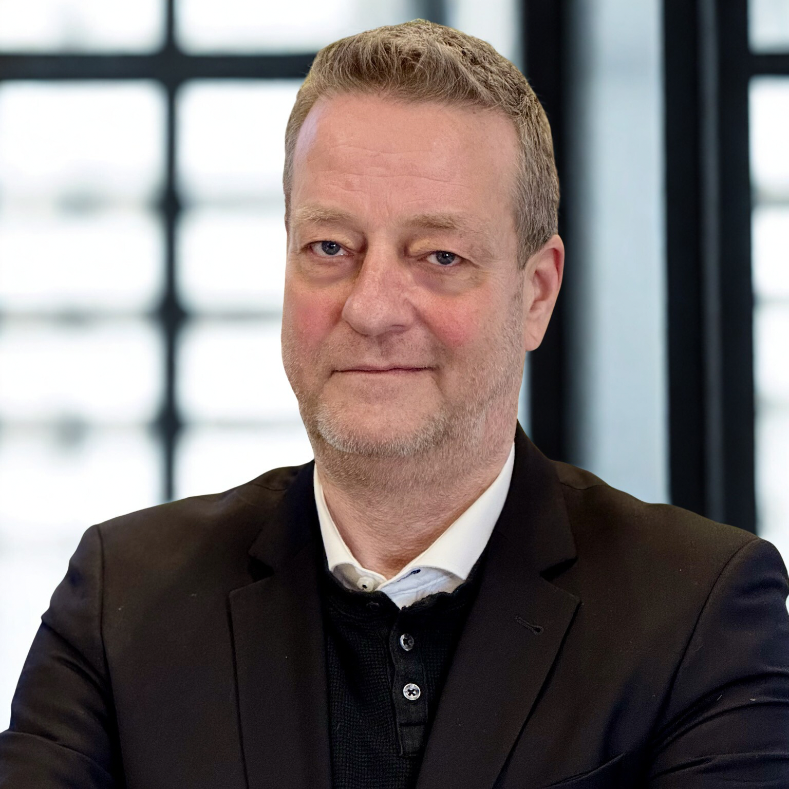 Mand i sort blazer med mørk trøje under og hvid skjorte, mellemblondt hår og gråligt skæg, New Yorker glasrude baggrund - Paul Rammeskow