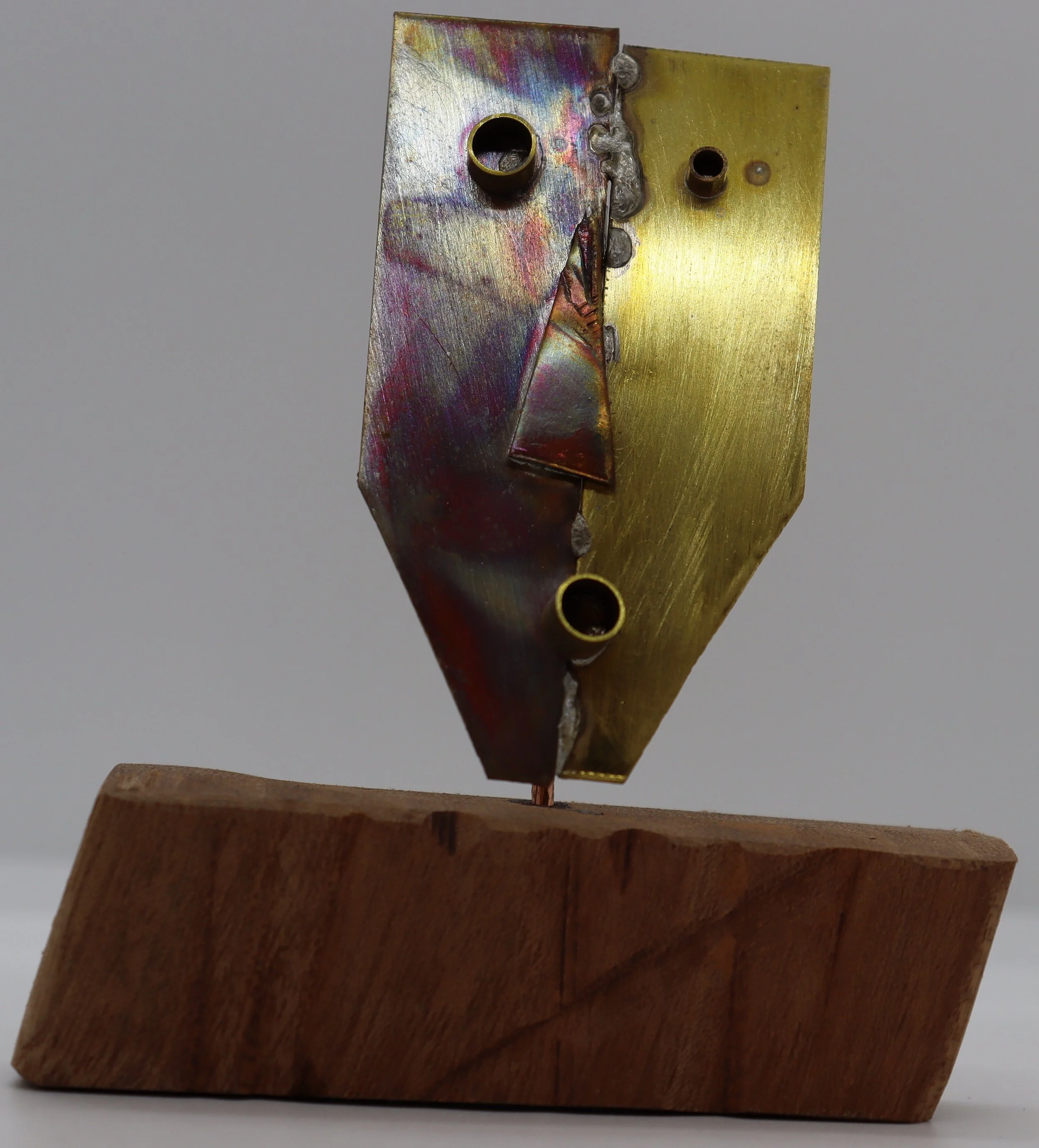 A small abstract sculpture with metallic and wooden elements. The sculpture features a geometric metal piece with a mix of gold, purple, and silver colors, mounted on a wooden base.