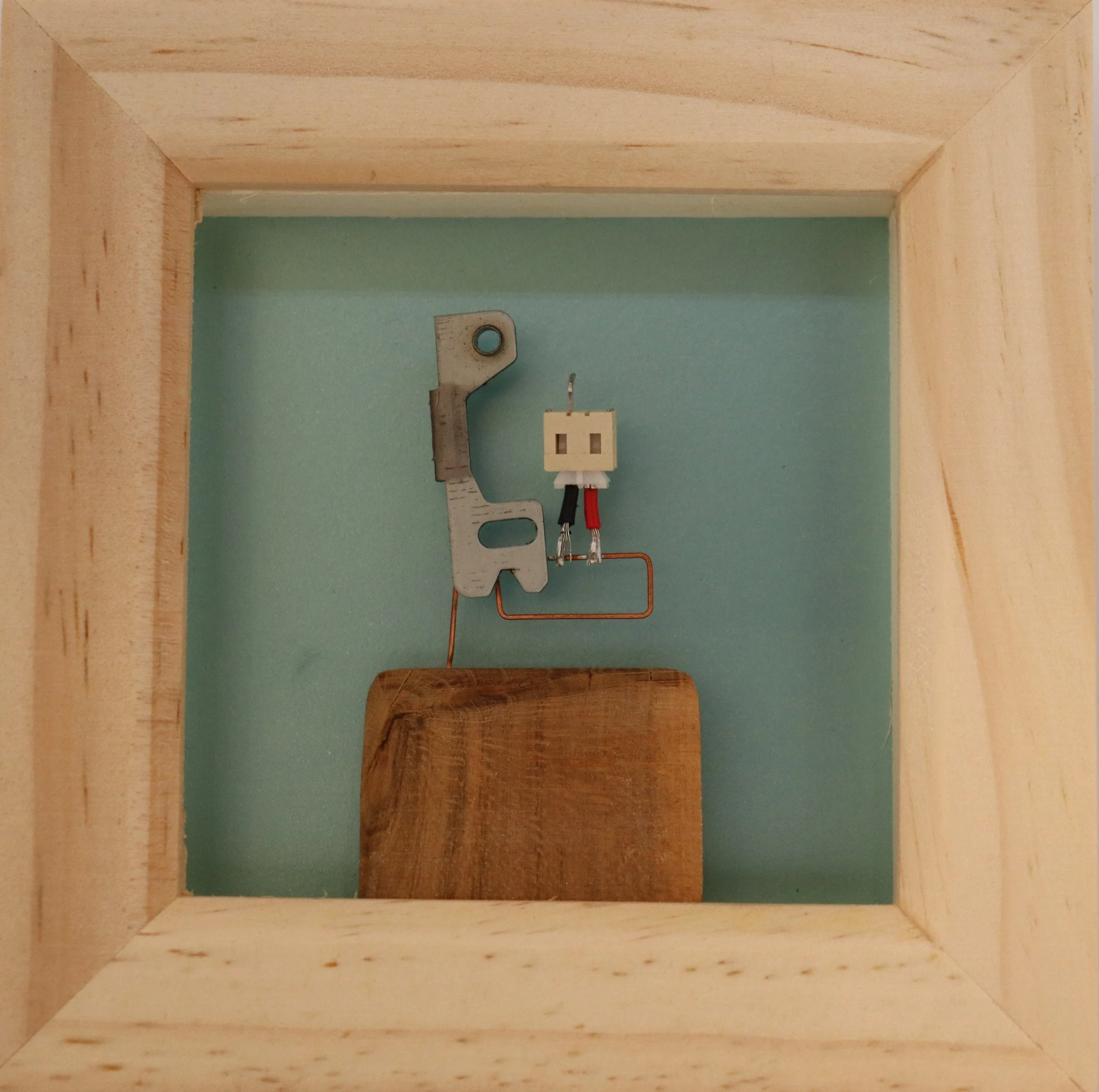 Inside a wooden frame, there are electrical components on a pastel green background, including a metal bracket, a small white socket with wires, and a wooden piece.