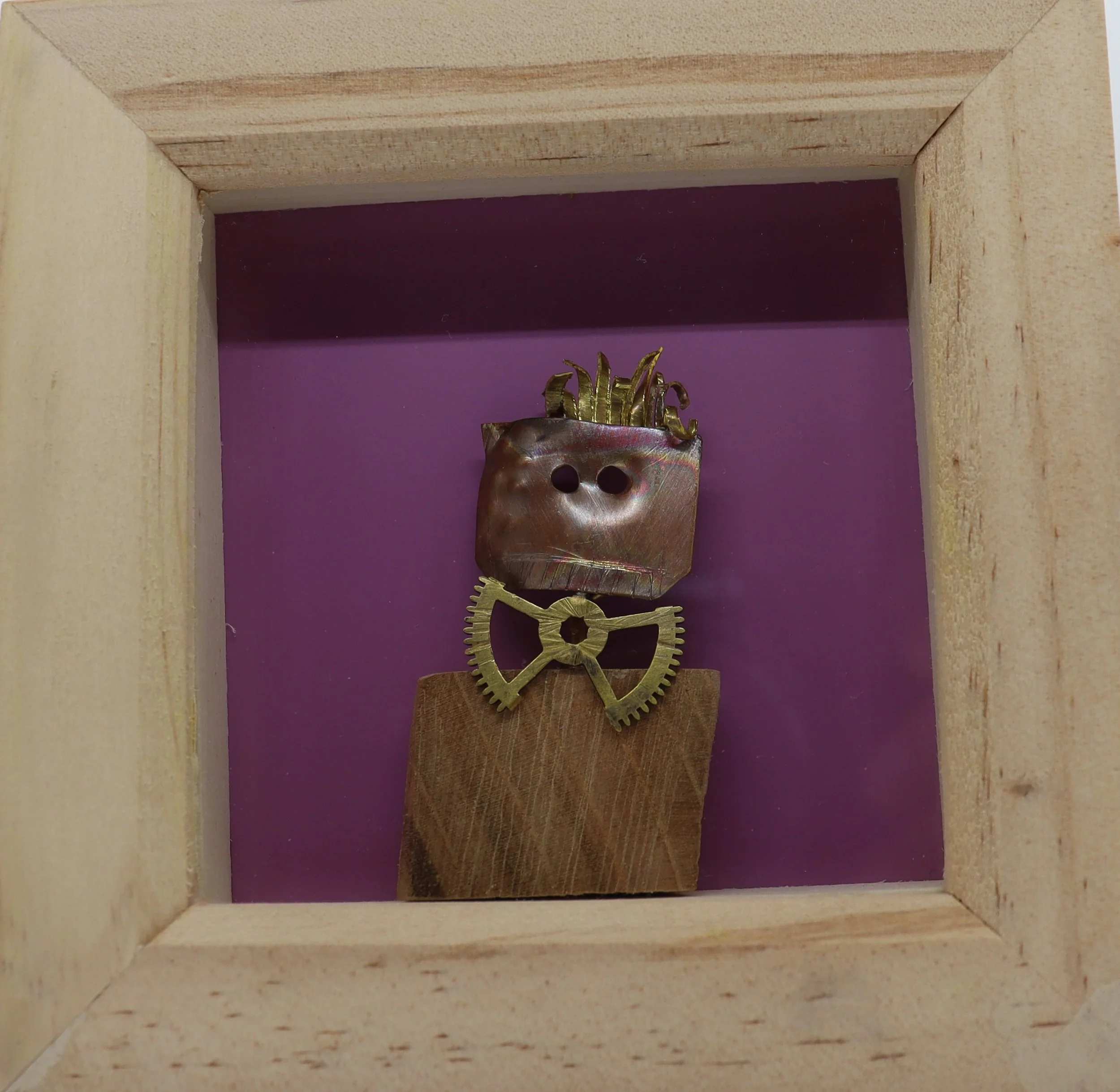 A mixed-media sculpture of a character with a square metallic face, two holes for eyes, spiky hair, a gear-shaped collar, and a rectangular body, displayed inside a wooden box with purple background.