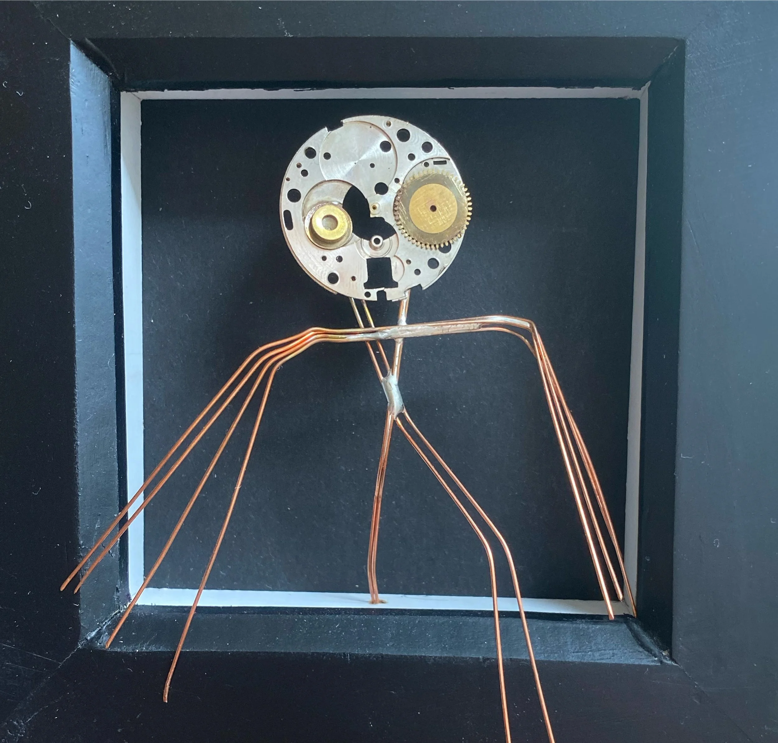 A mechanical humanoid figure made with clock parts, copper wires forming limbs, with a clock face as the head, inside a black frame.